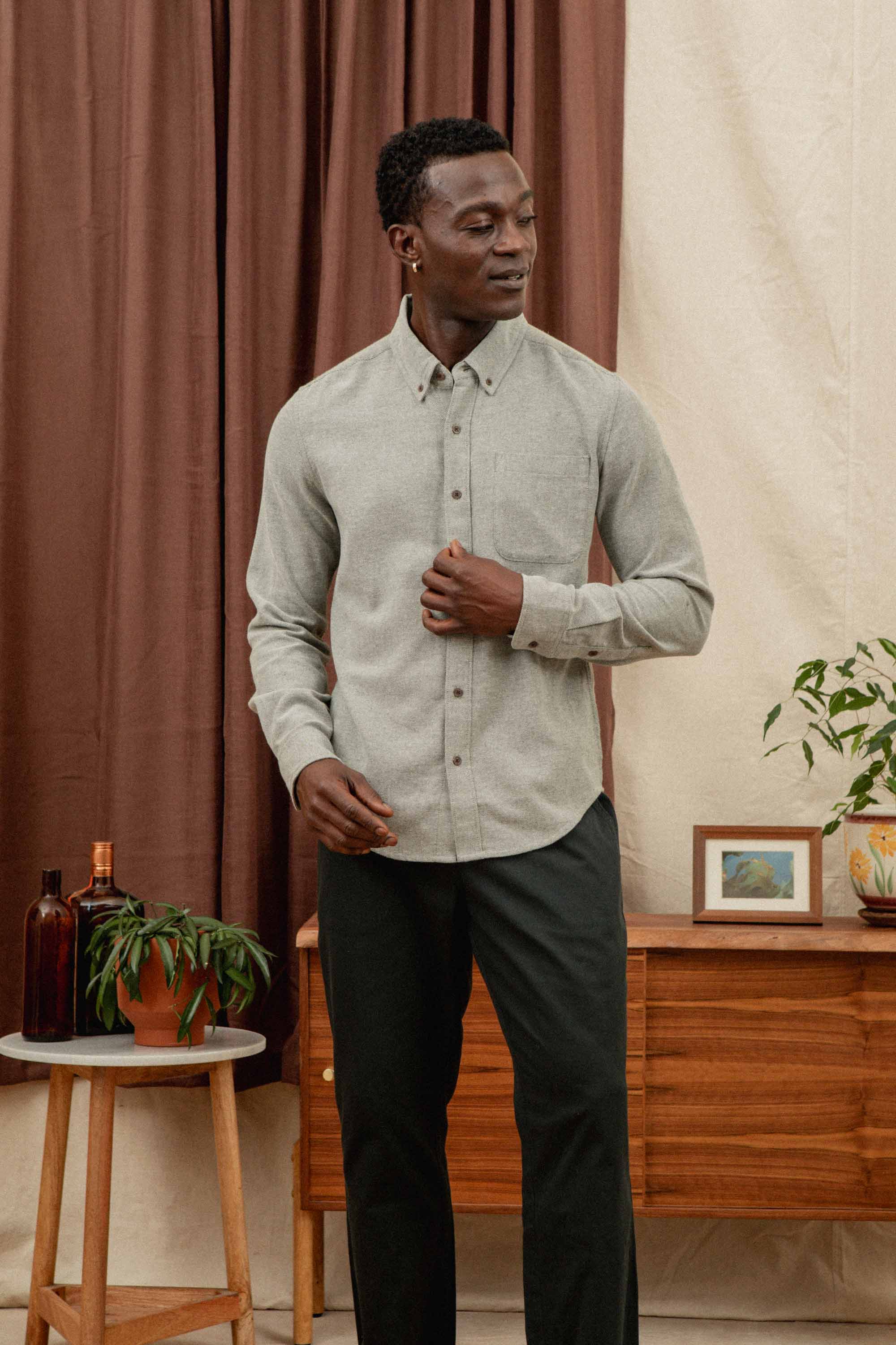A man wearing the Bridge & Burn Sutton Slim Shirt in olive twill and black pants stands indoors near a wooden table with plants, bottles, framed art, and brown and beige curtains in the background.