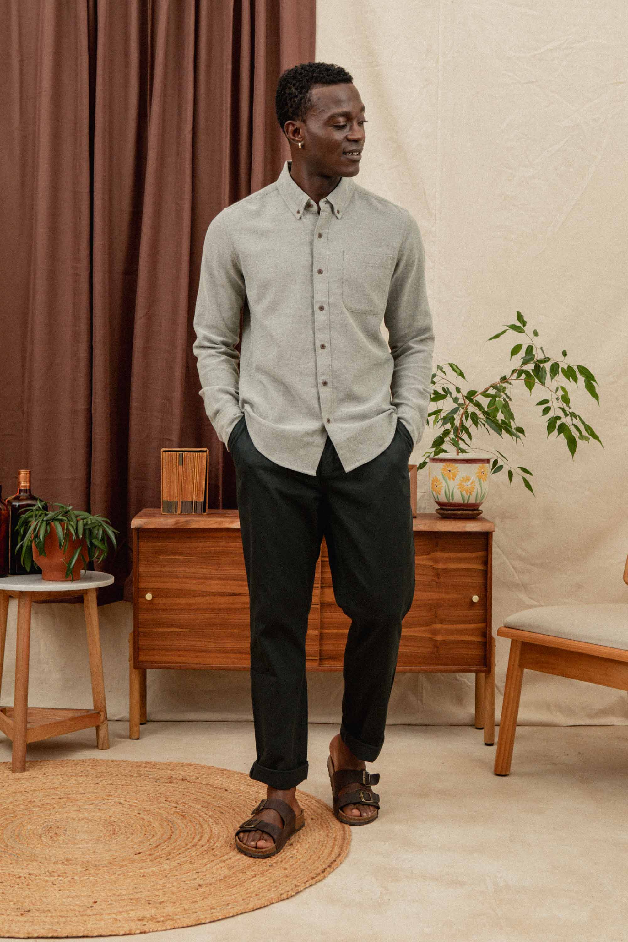 A man wears the Bridge & Burn Sutton Slim Shirt in olive twill, paired with black pants and sandals. He stands indoors among wooden furniture, potted plants, brown curtains, and a round rug.