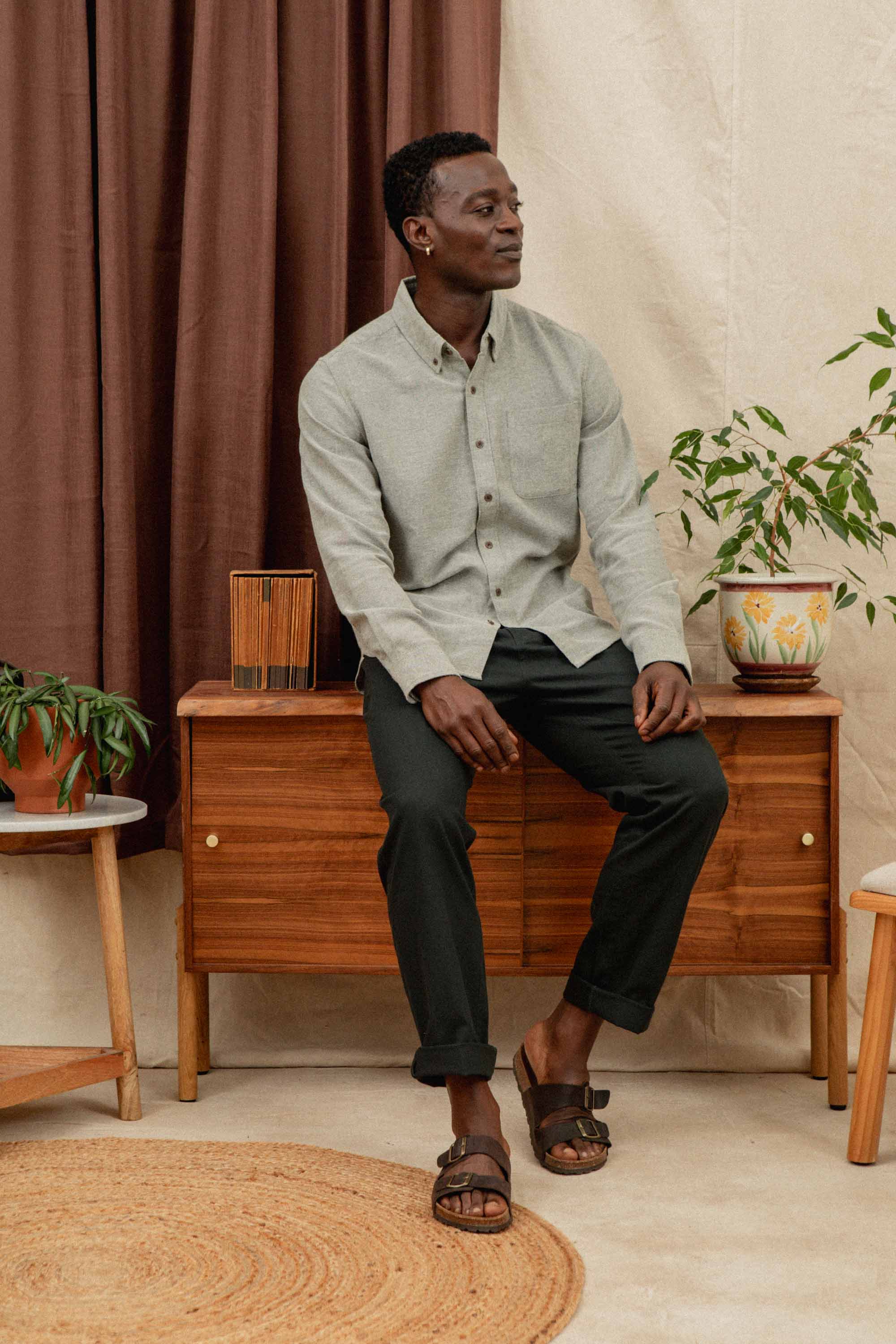 A man wears the Bridge & Burn Sutton Slim Shirt in olive twill and dark pants, seated on a wooden cabinet with potted plants, round woven rug beneath him, and brown-beige curtains in the background.