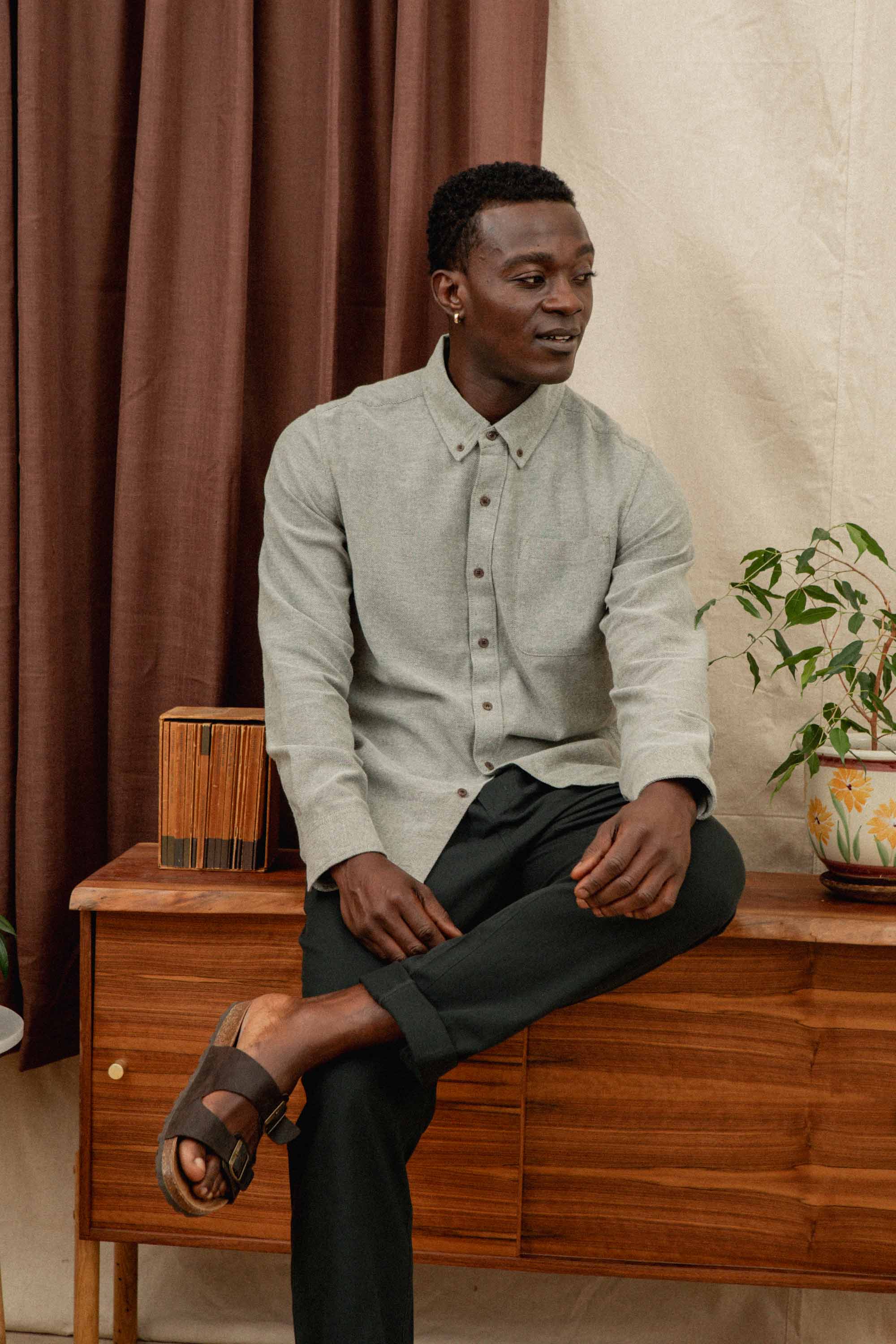 A man in a Bridge & Burn Sutton Slim Shirt in Olive Twill, dark pants, and brown sandals sits on a wooden sideboard beside a potted plant and wooden decor, set against brown curtains and a cream wall.