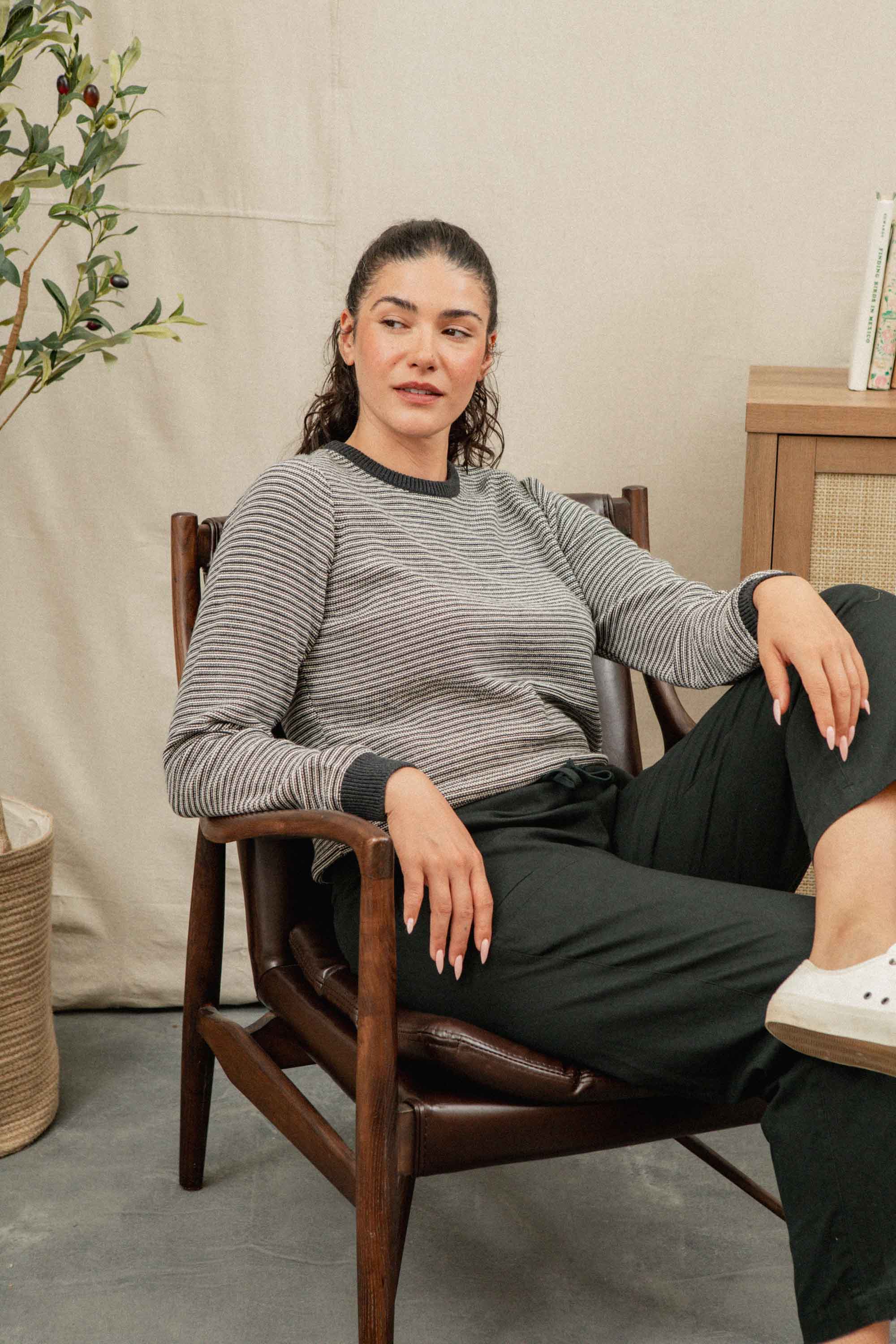 A woman with curly hair relaxes in a wooden chair, wearing the Ansel Sweater in Charcoal Stripe by Bridge & Burn—crafted from recycled fibers—paired with black pants and white sneakers in a cozy, neutral-toned room.