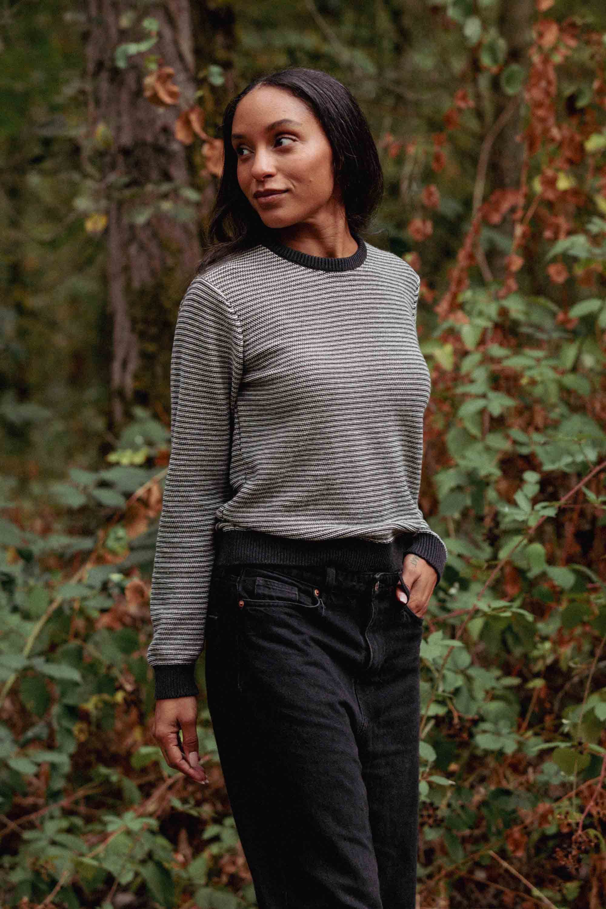 A woman with long dark hair stands outdoors among green foliage, wearing the Bridge & Burn Ansel Sweater in Charcoal Stripe and black jeans, smiling softly while looking to the side with one hand in her pocket.