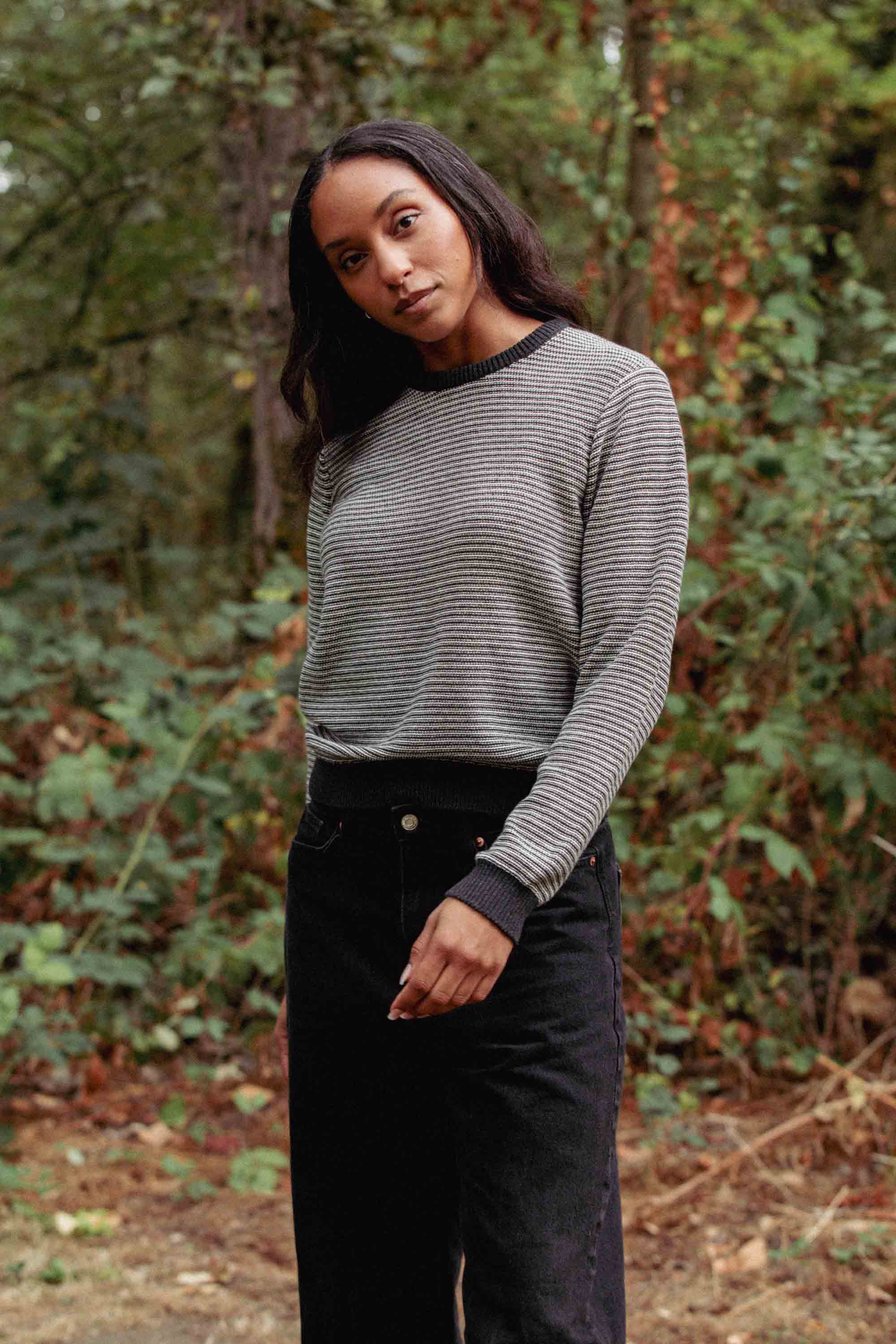 A woman with long dark hair stands outdoors in front of greenery, wearing the Bridge & Burn Ansel Sweater in Charcoal Stripe and black pants, her head slightly tilted and one arm relaxed by her side.