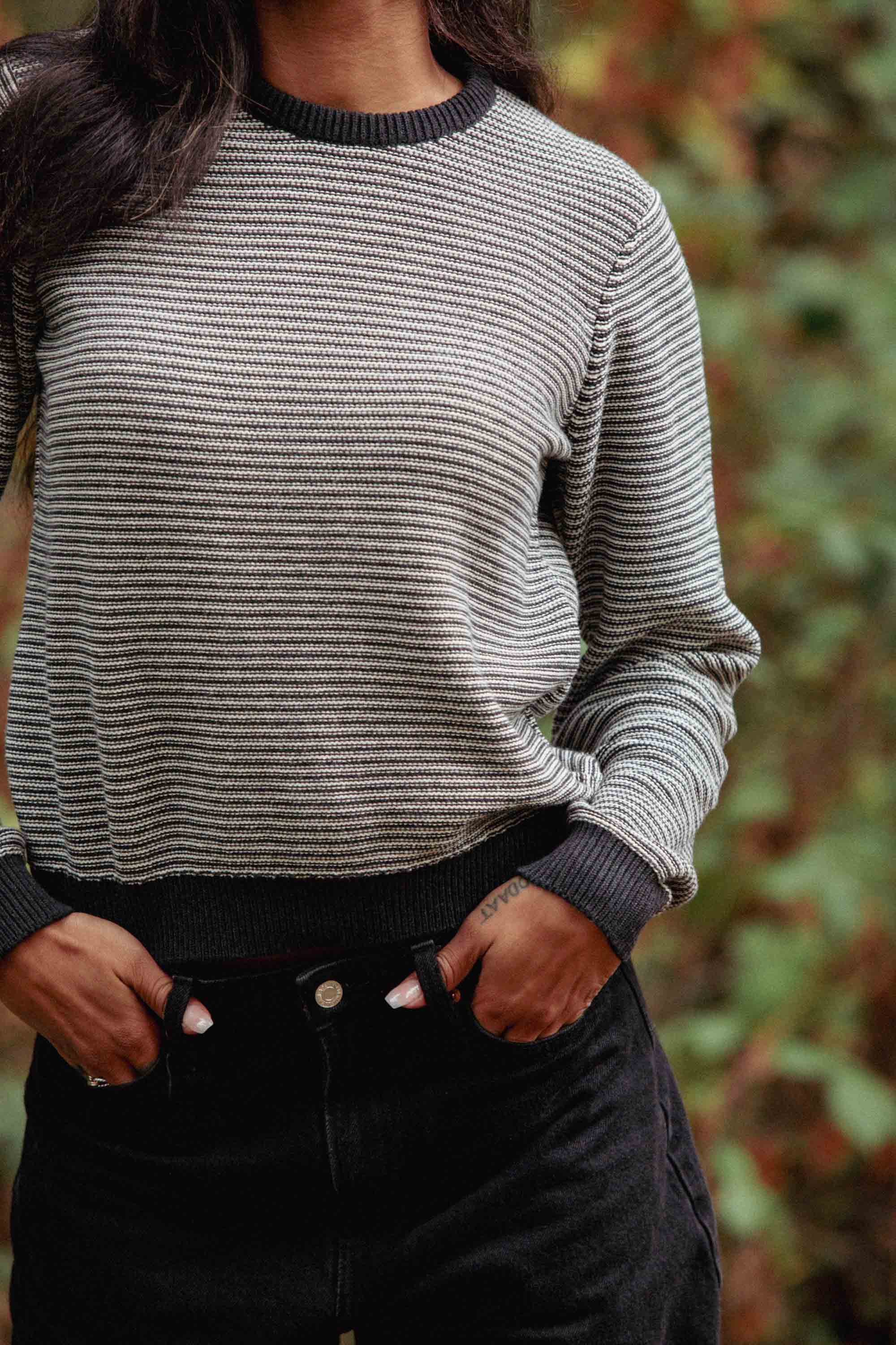 A person stands outdoors in the Bridge & Burn Ansel Sweater in Charcoal Stripe—crafted from recycled fibers—and black pants, hands in pockets. Blurred green foliage highlights this sustainable essential.
