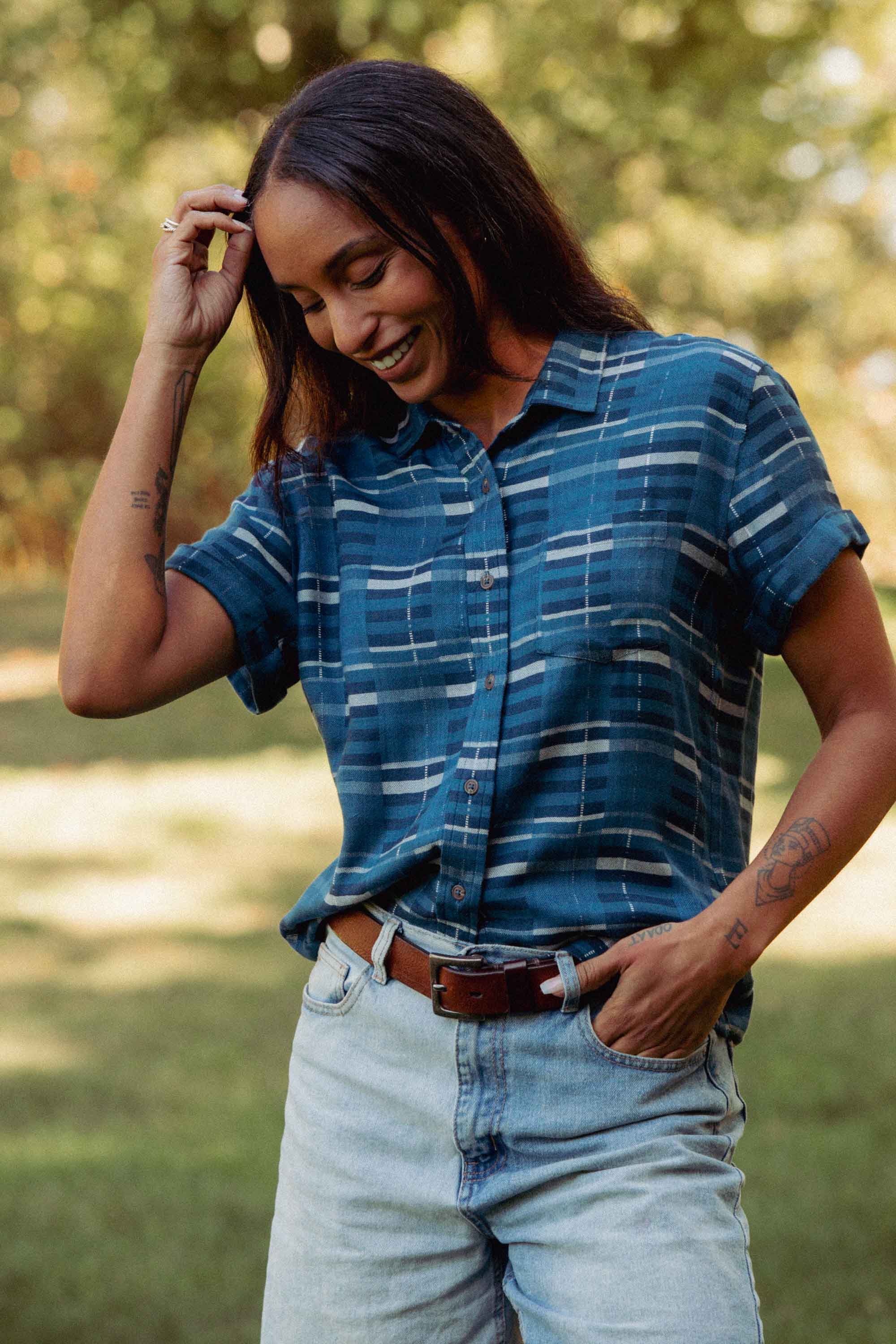 A woman stands on grass, eyes closed and smiling, wearing the Bridge & Burn Bea Shirt in Stormcrest Stripe with light jeans and a brown belt. Sunlight filters through green trees, highlighting her arm tattoos.