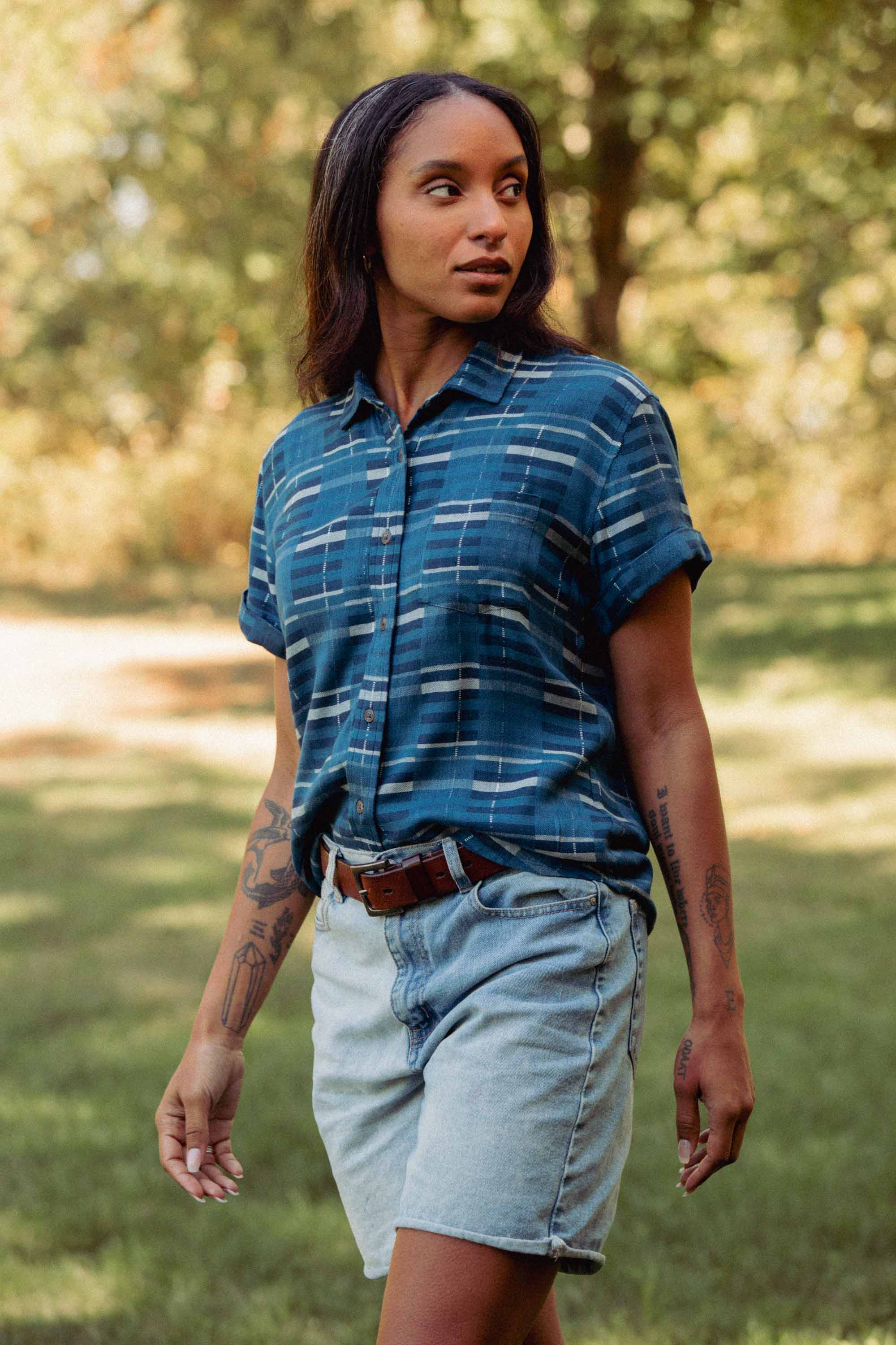 A person with long dark hair wears the Bridge & Burn Bea Shirt in Stormcrest Stripe, tucked into light denim shorts with a brown belt, standing outdoors on grass with blurred trees in the background.