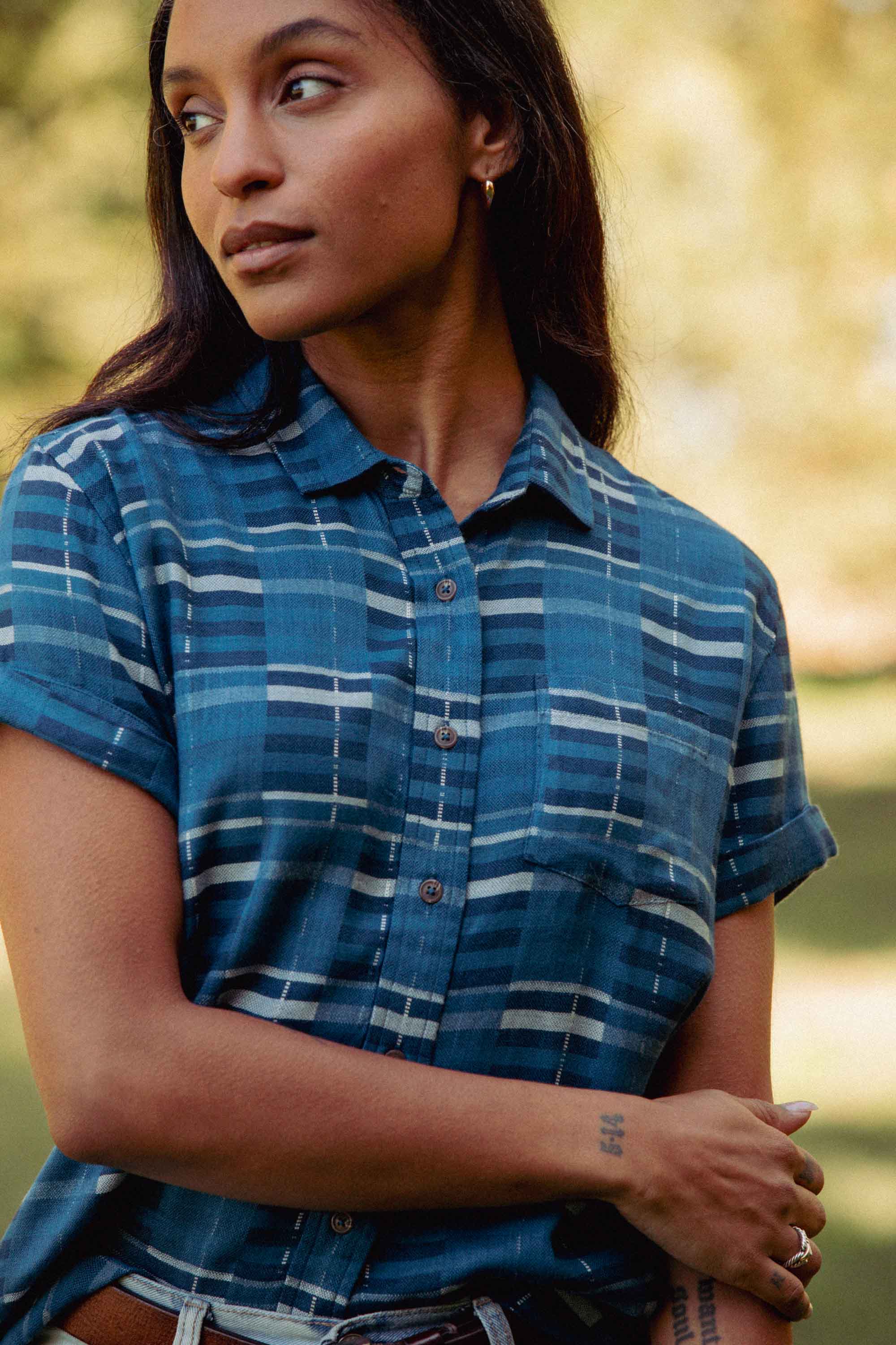 A person with long dark hair wears the Bridge & Burn Bea Shirt in Stormcrest Stripe—a tailored fit, short-sleeve button-down—while standing outdoors amid blurred trees, looking off to the side with a calm expression.