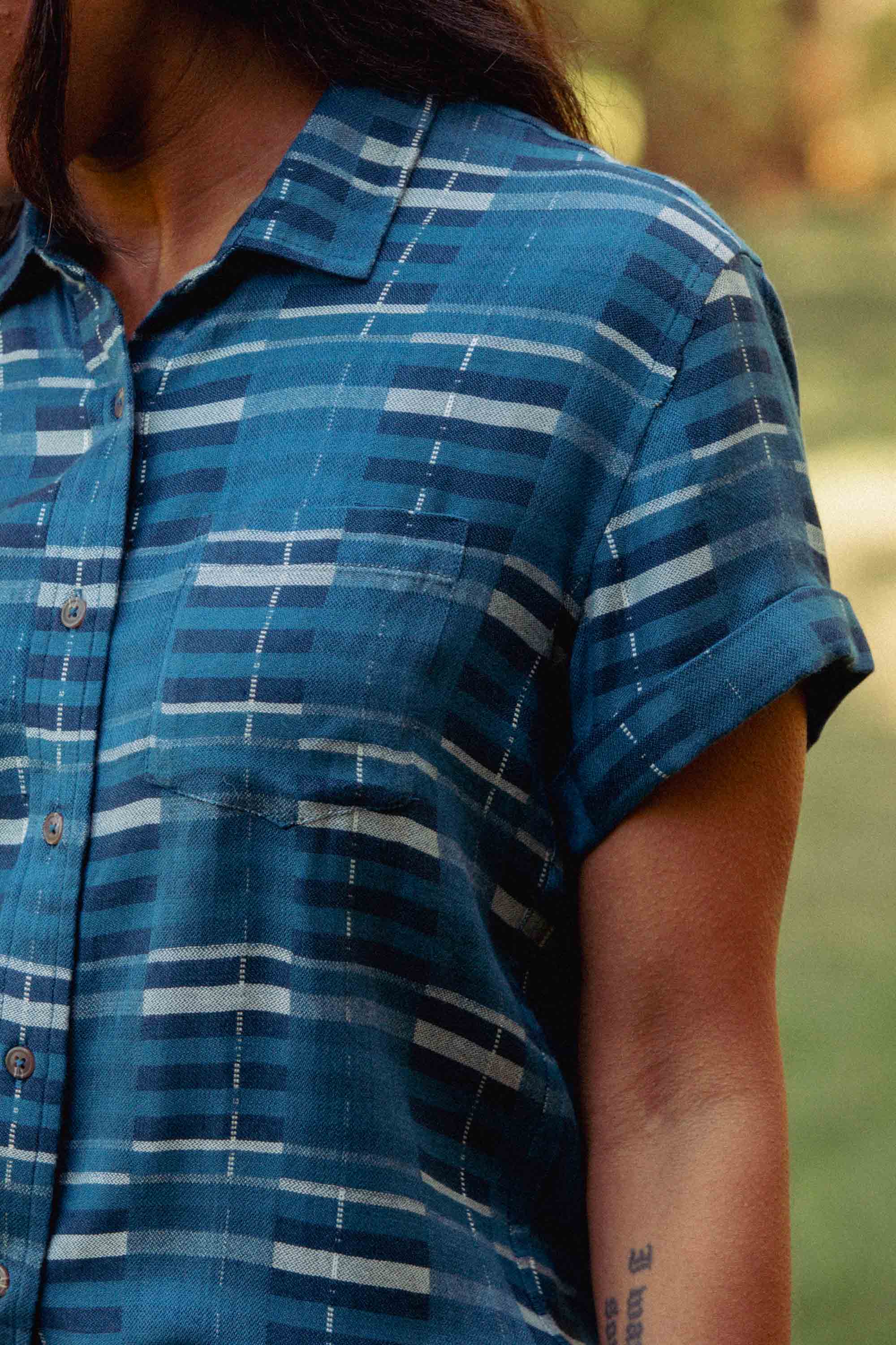 A person outdoors wears the Bridge & Burn Bea Shirt in Stormcrest Stripe, a blue plaid short-sleeve button-down. Their tattooed left arm is visible against blurred greenery.