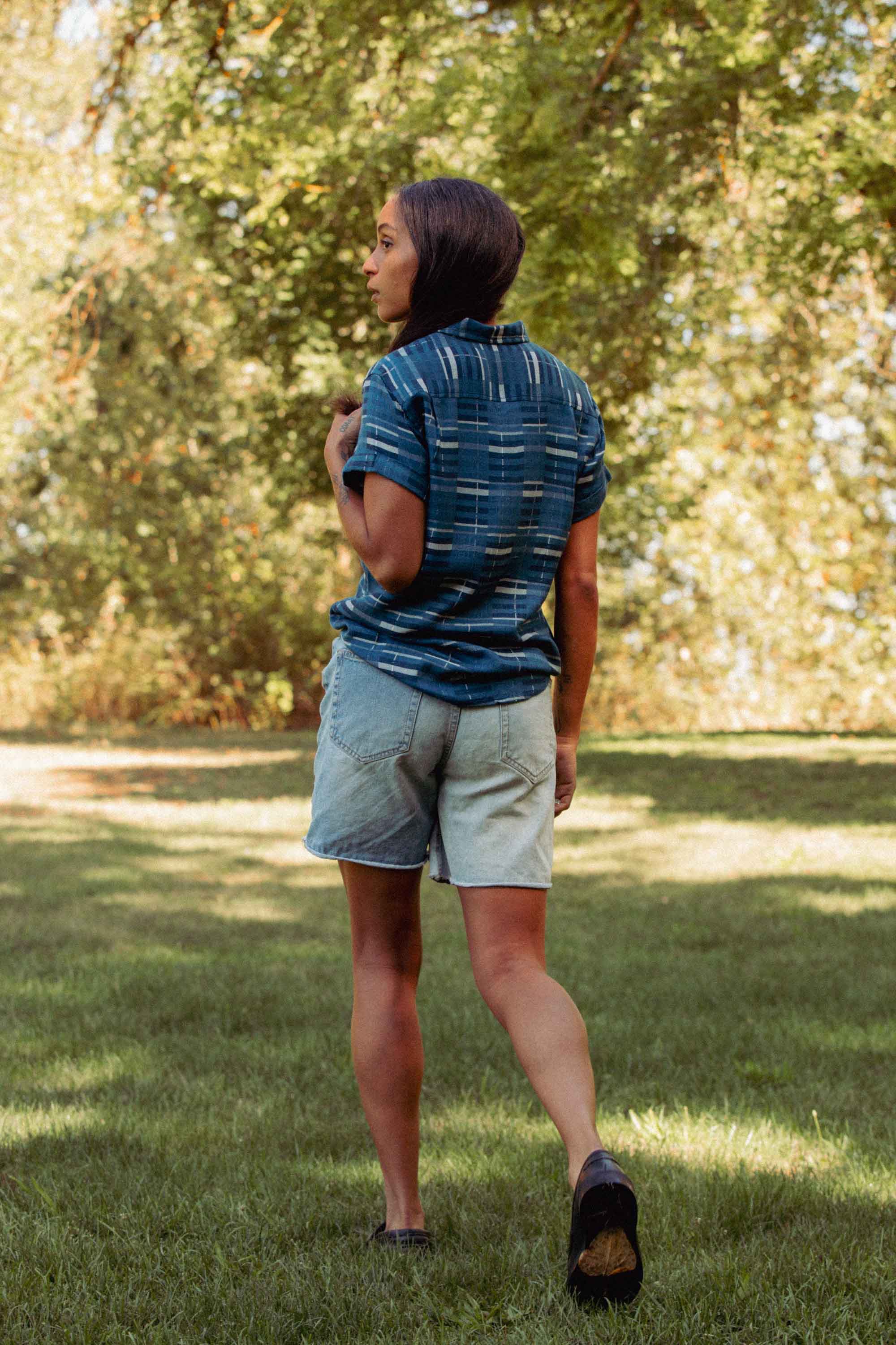 Wearing the Bridge & Burn Bea Shirt in Stormcrest Stripe and denim shorts, a person walks on grass in a sunlit park, glancing over their shoulder with lush green trees in the background.