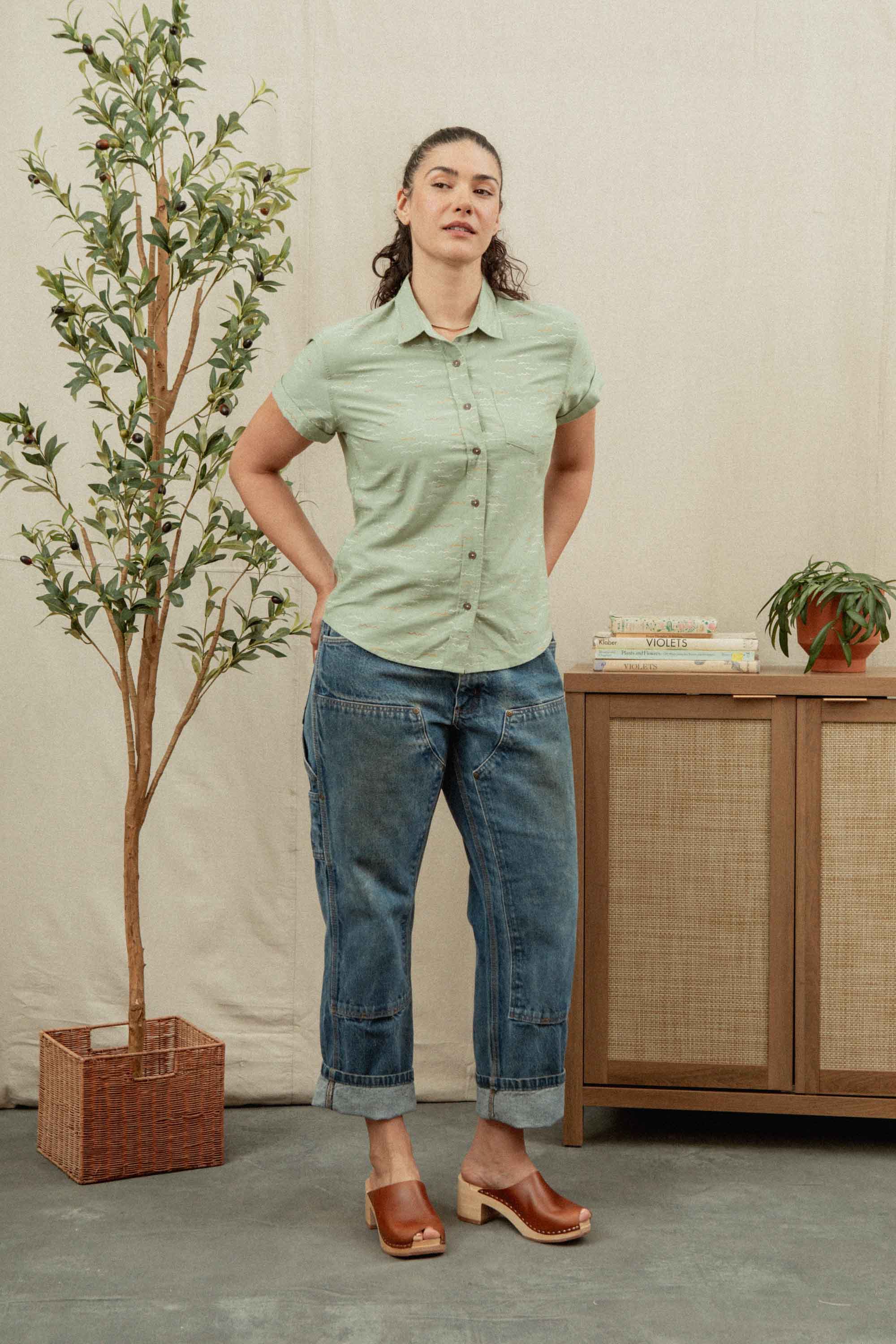 A woman stands indoors wearing the Bridge & Burn Bea Shirt in Teal Tide Waves, paired with cuffed blue jeans and brown clogs. She is by a leafy potted plant and a wooden cabinet with books and a small plant on top.