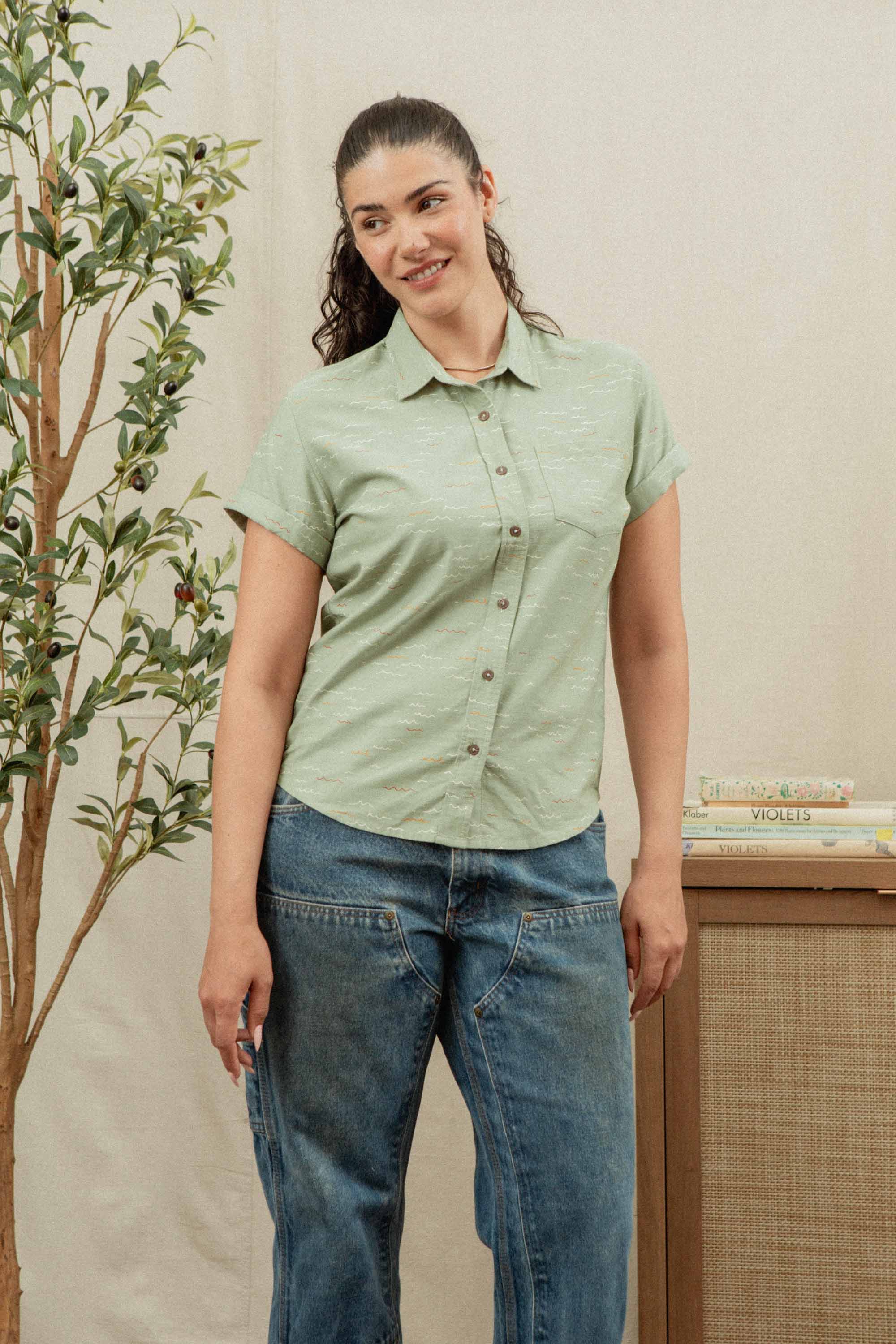 A woman with dark hair in a ponytail wears the Bridge & Burn Bea Shirt in Teal Tide Waves and loose blue jeans, standing indoors near a leafy plant and a wooden table with stacked books.