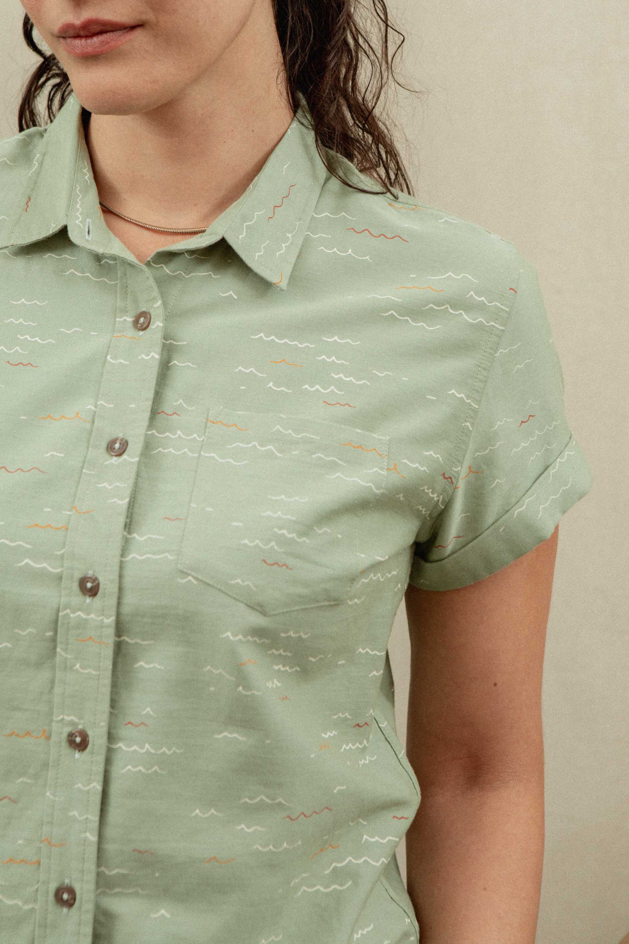 A woman wears the Bridge & Burn Bea Shirt in Teal Tide Waves—an organic cotton, light green short-sleeve button-down with a subtle wavy pattern. Her partially visible face and wavy hair are set against a plain light beige background.