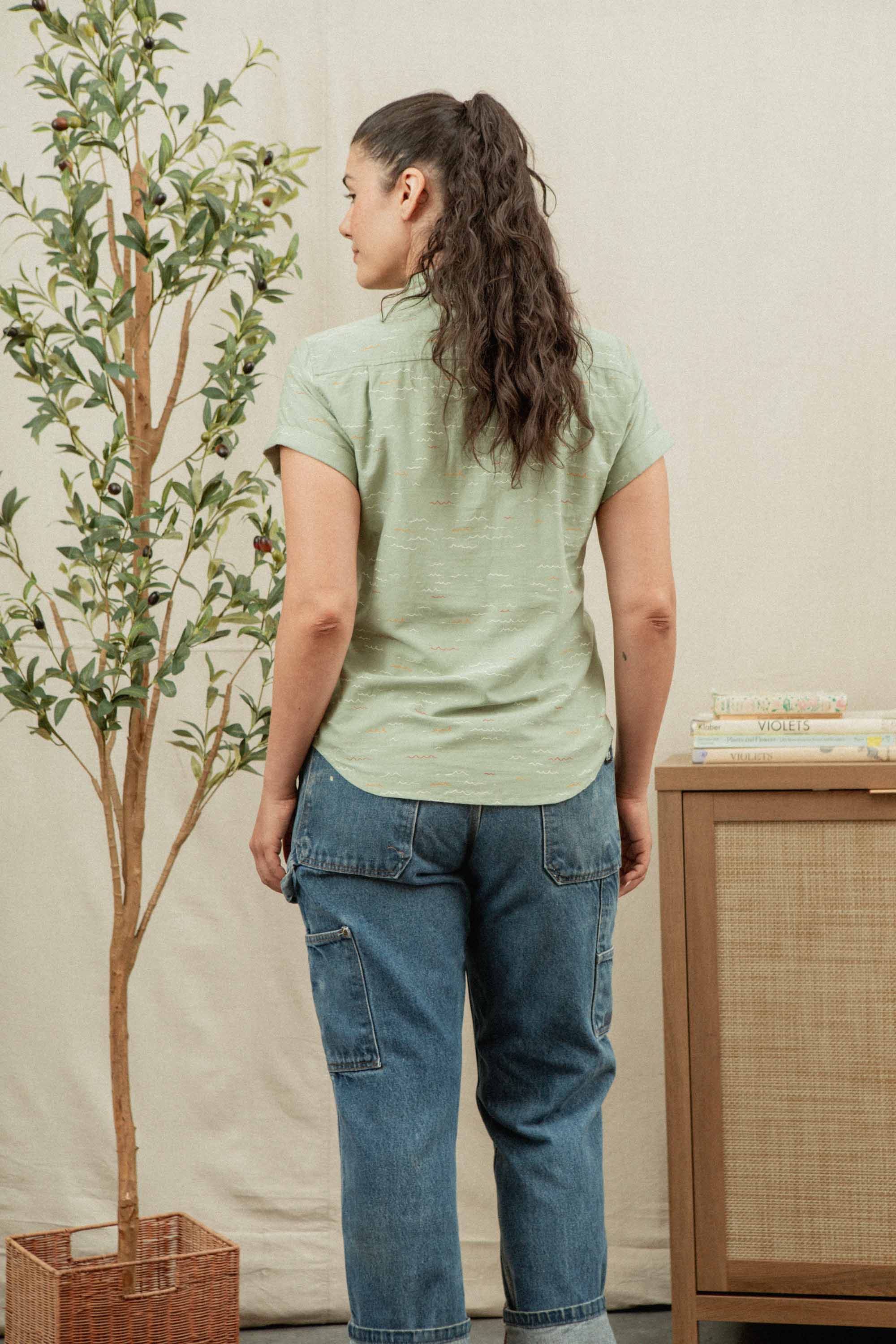 A woman with long, wavy brown hair in a ponytail stands facing away, wearing the Bridge & Burn Bea Shirt in Teal Tide Waves and blue jeans. Beside her are a potted tree and a wooden cabinet with books.