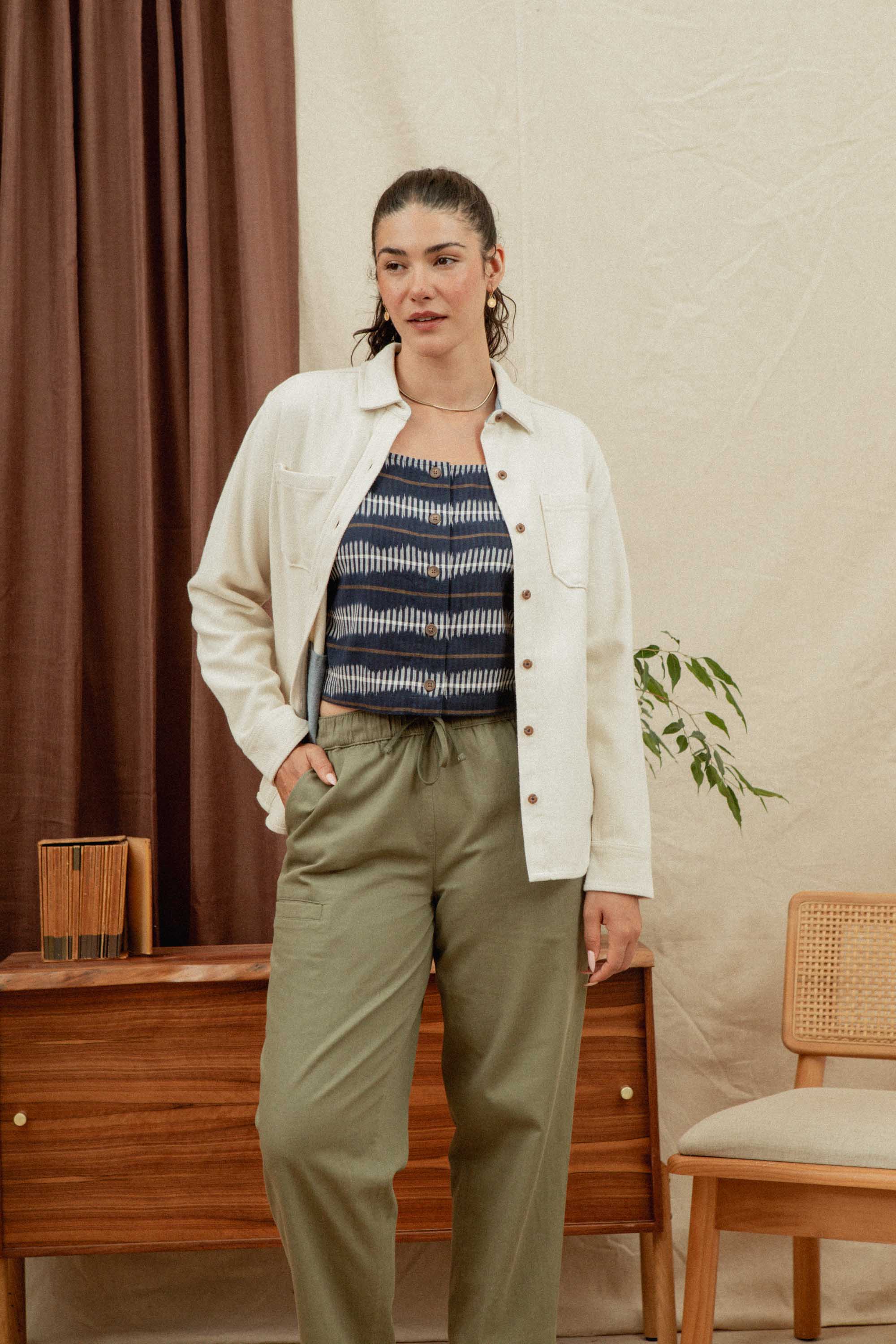 A woman stands indoors wearing the Bridge & Burn Dawson Overshirt in Ivory Twill over a blue patterned top and olive green pants, hand in her pocket, with neutral decor and a plant in the background.