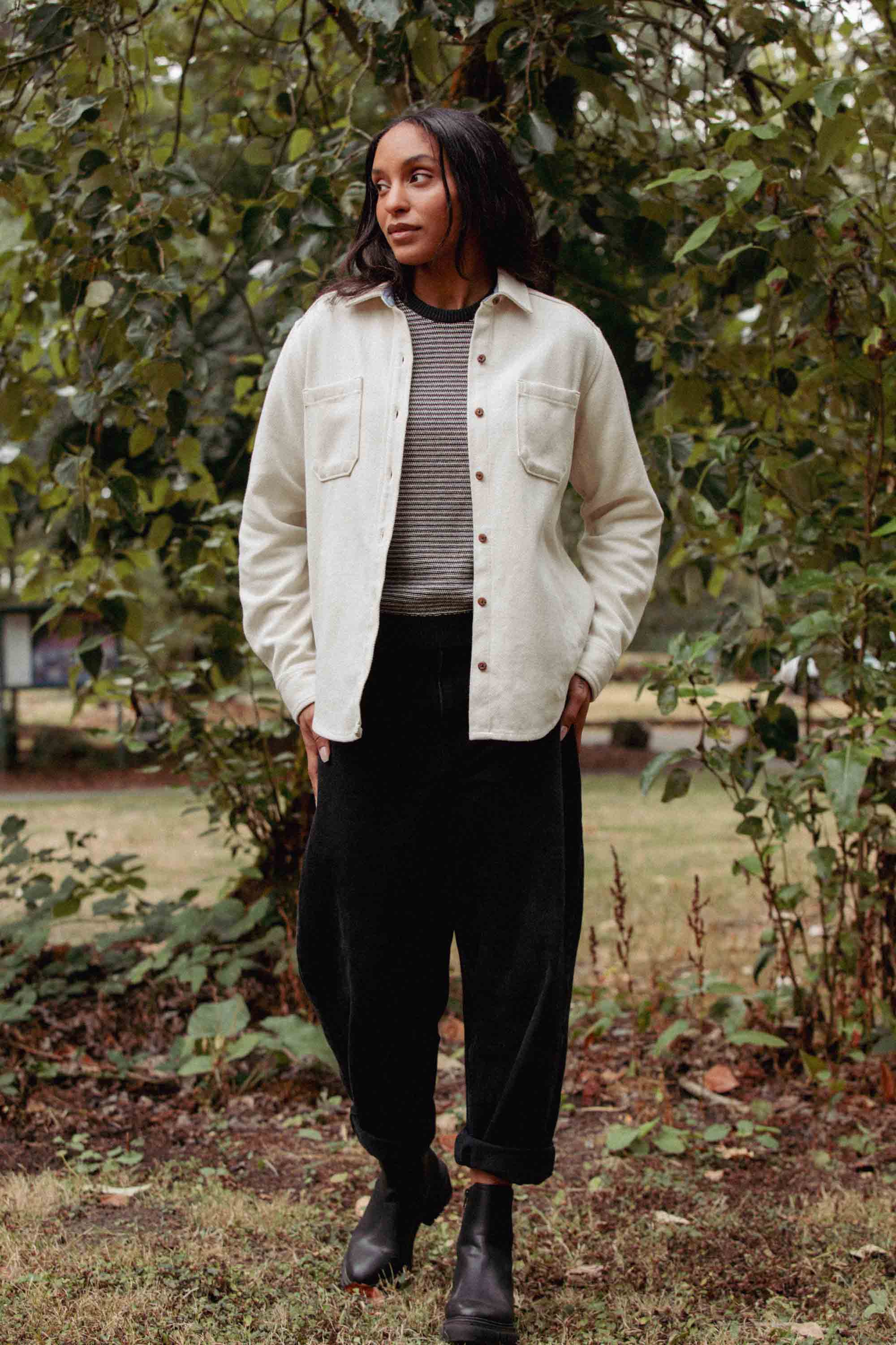Standing outdoors before green foliage, a person wears the Bridge & Burn Dawson Overshirt in Ivory Twill over a striped top, paired with black pants and boots, hands in pockets and looking off to the side.