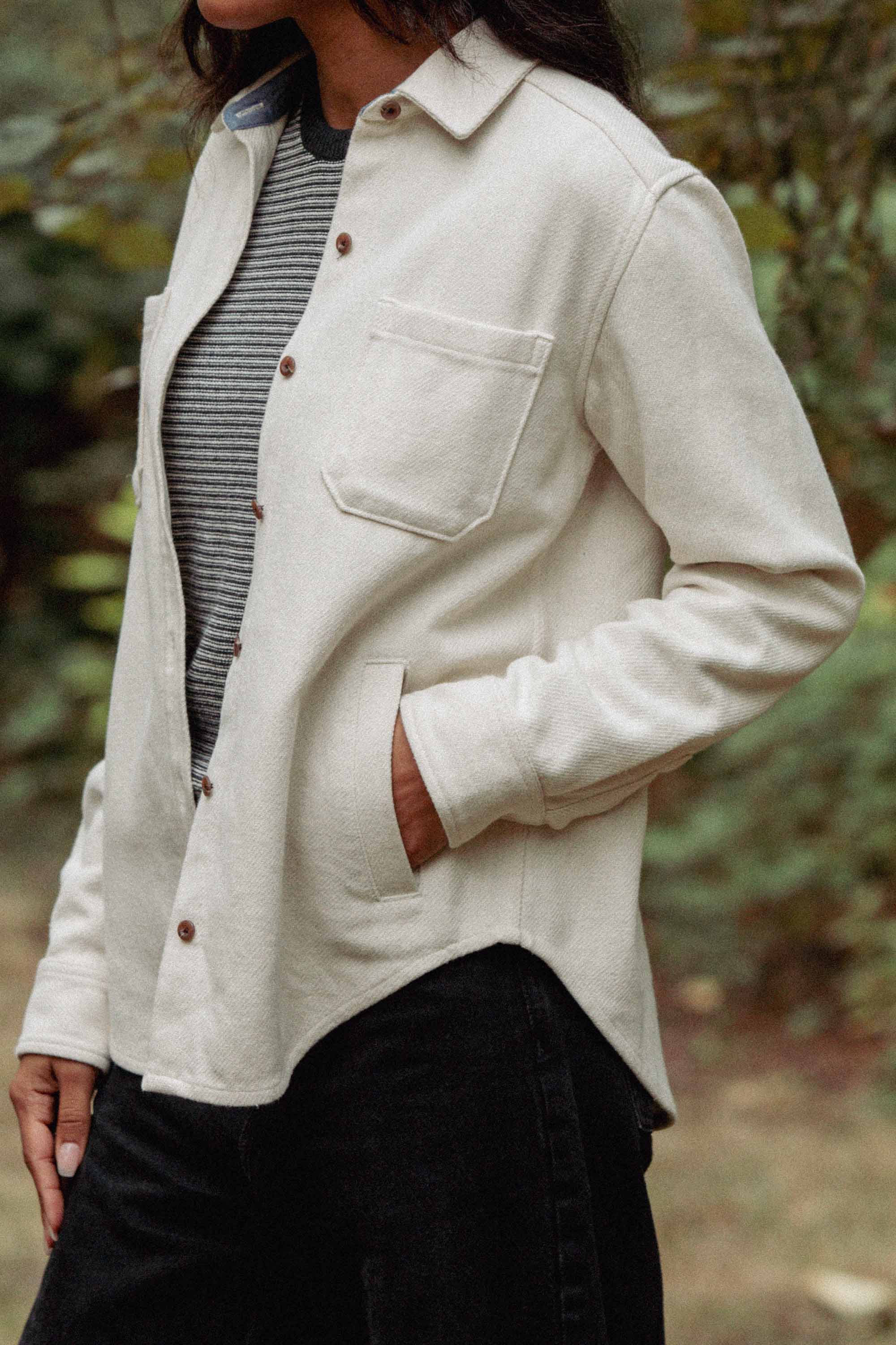 A person stands outdoors wearing the Bridge & Burn Dawson Overshirt in Ivory Twill over a black and white striped top, with one hand in their black pants pocket. Blurred greenery in the background highlights their sustainable style.