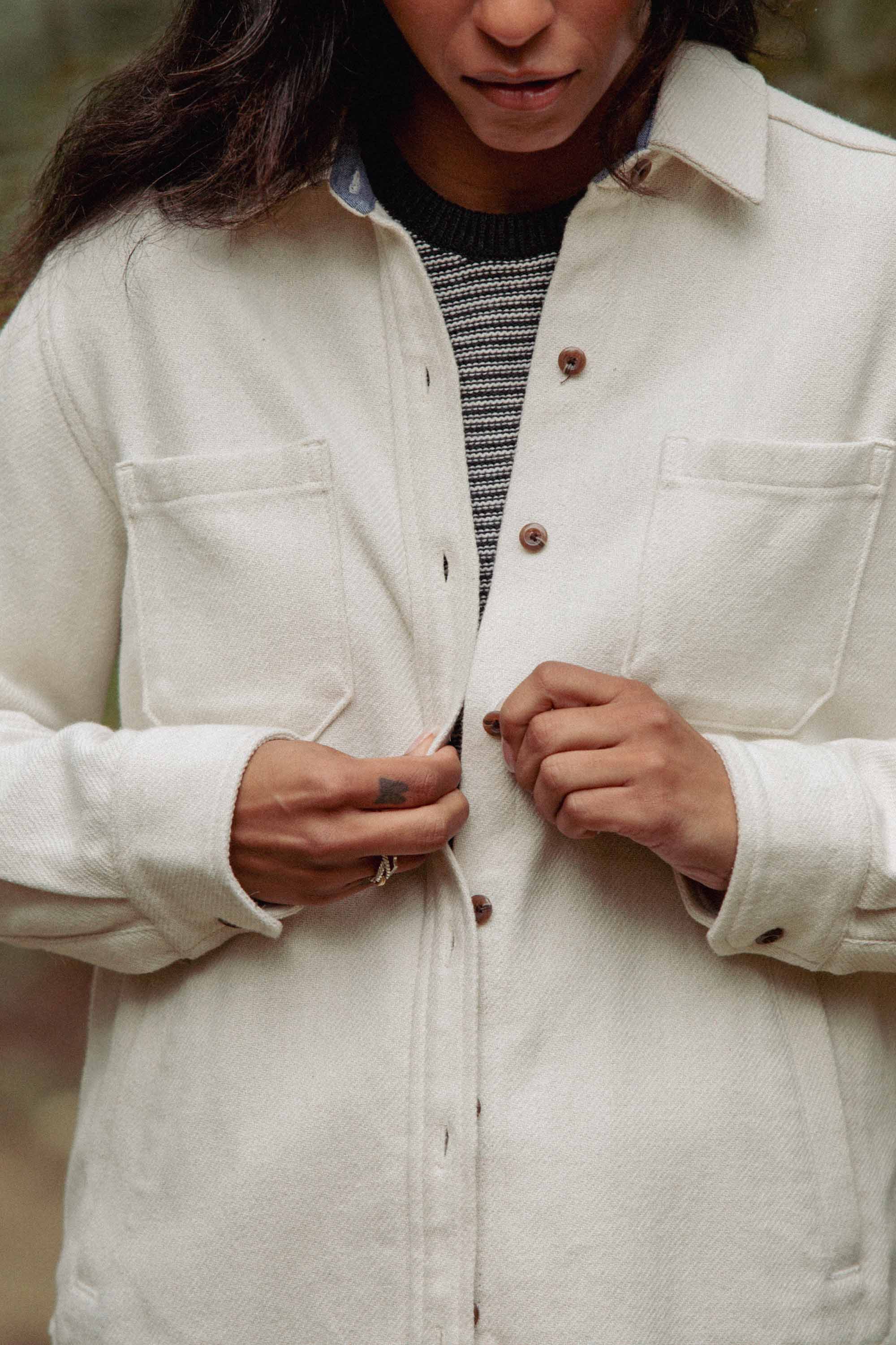 Buttoning the Dawson Overshirt in Ivory Twill by Bridge & Burn over a black-and-white striped top, only the lower half of the person’s face and torso are visible, drawing focus to this sustainable style choice.