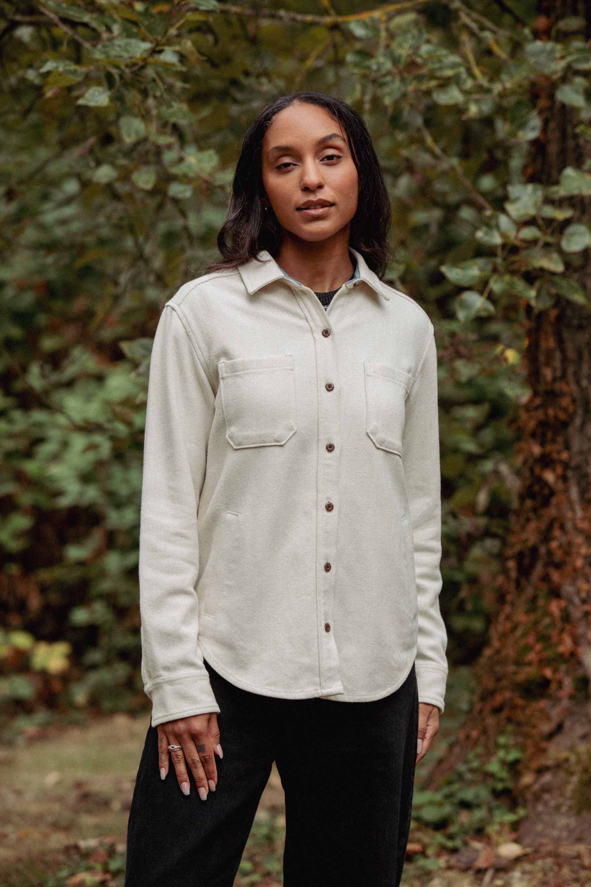 A woman stands outdoors before green foliage, wearing the Dawson Overshirt in Ivory Twill by Bridge & Burn with two chest pockets and black pants. She looks at the camera, showcasing stylish sustainable fashion.