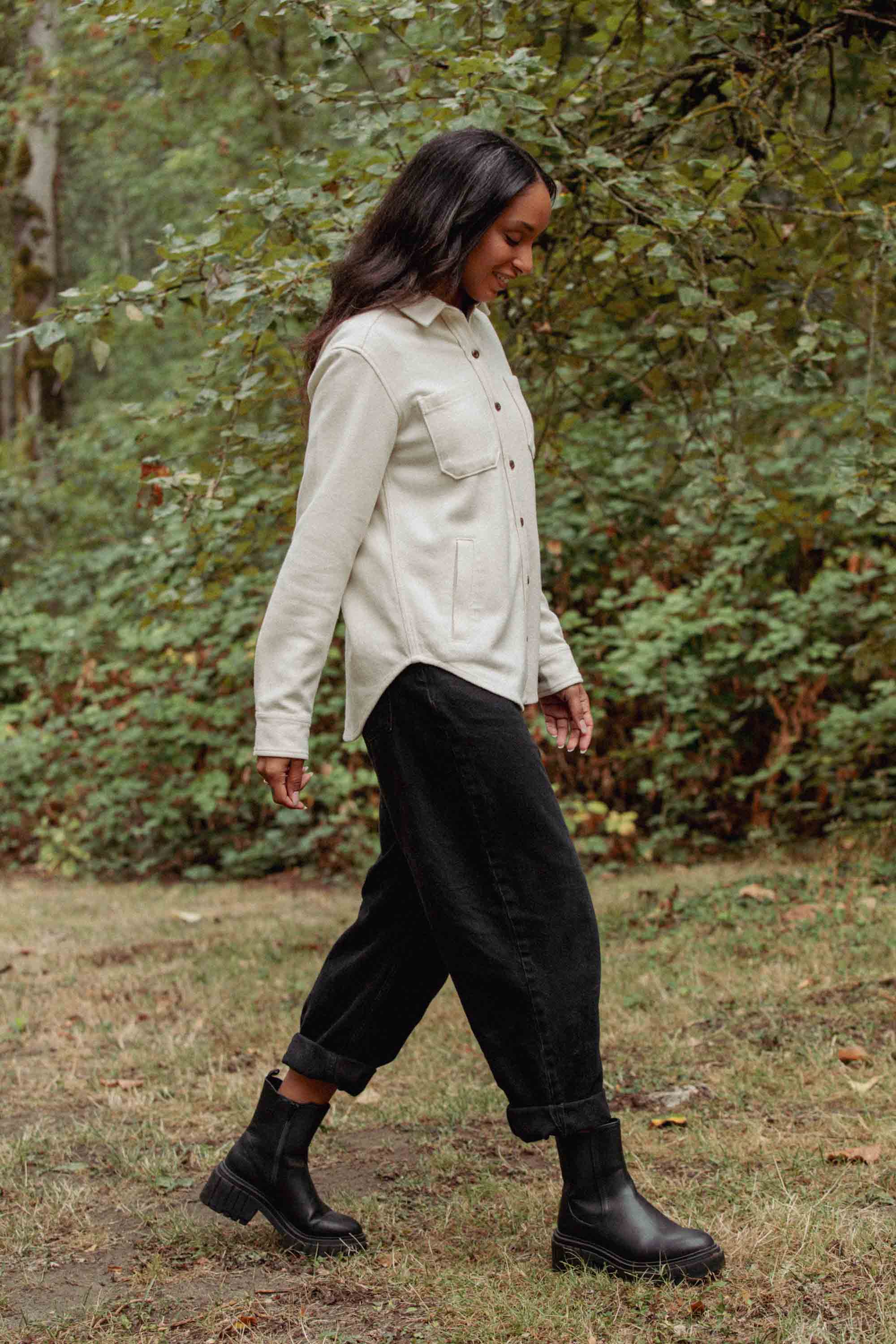 A person walks on grass wearing the Bridge & Burn Dawson Overshirt in Ivory Twill, paired with black pants and black ankle boots, with trees and greenery in the background.