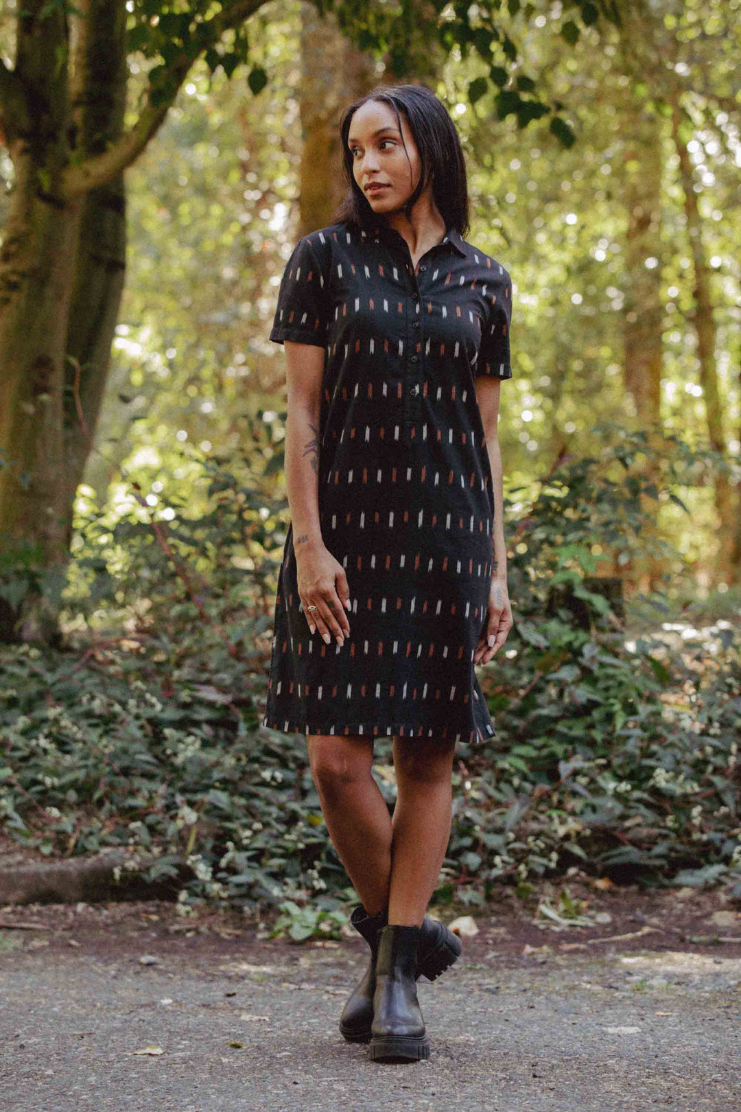 A woman stands outdoors in sunlight, wearing the Bridge & Burn Delia Dress in Dotted Horizon—an organic cotton shirt dress with a colorful ikat-inspired pattern—and black boots. She has straight dark hair and looks to the side, appearing relaxed.