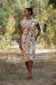 On a dirt path surrounded by greenery, a woman in a bun stands with one hand in her pocket, gazing sideways. She wears Bridge & Burn’s Eloise Dress in Pop Leaf—a neutral-toned A-line essential for any wardrobe.