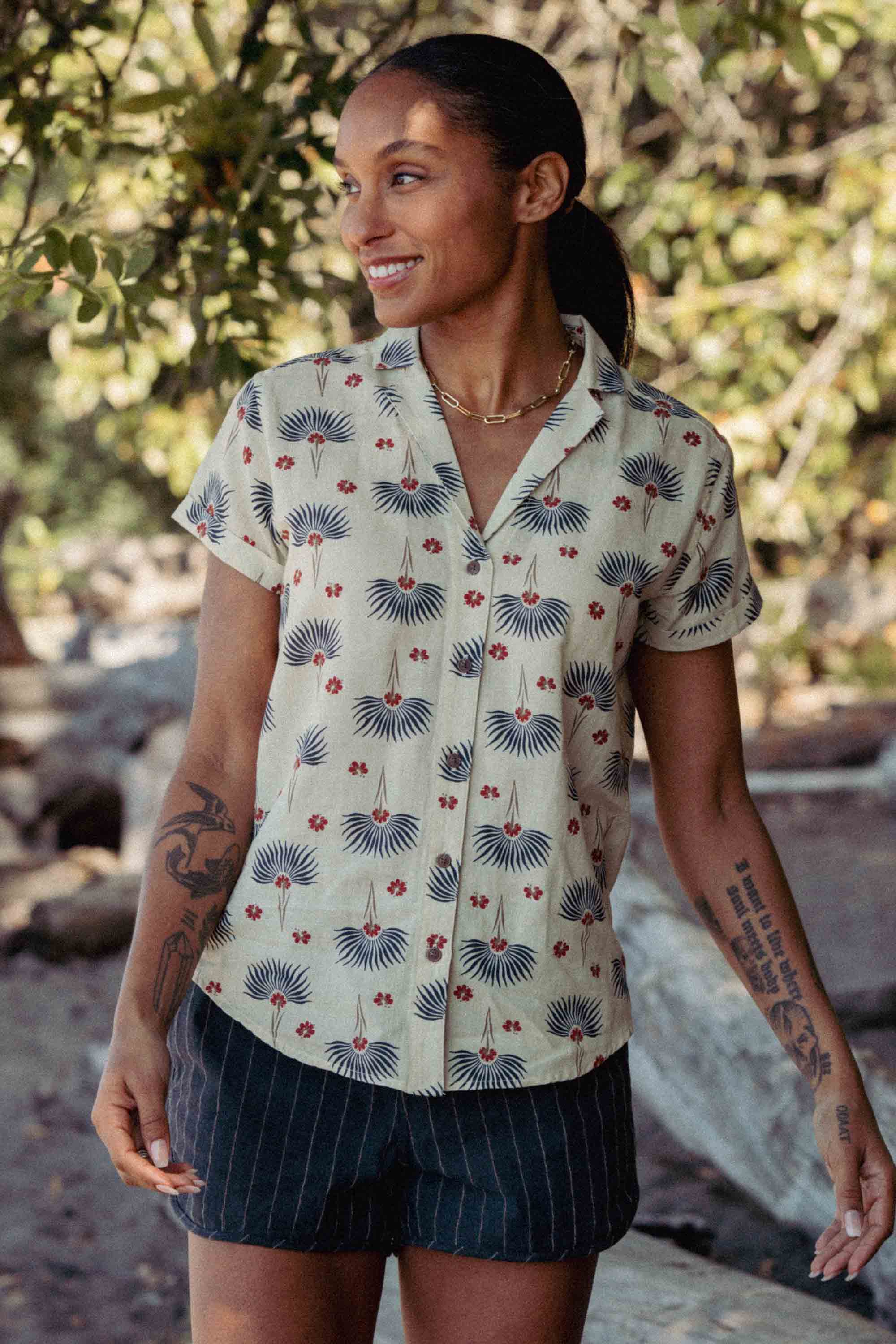 Wearing the Bridge & Burn Innes Shirt in Aloha Floral, a person stands outdoors smiling and looking to the side, pairing the relaxed fit button-up with striped shorts. Trees and rocks can be seen in the background.
