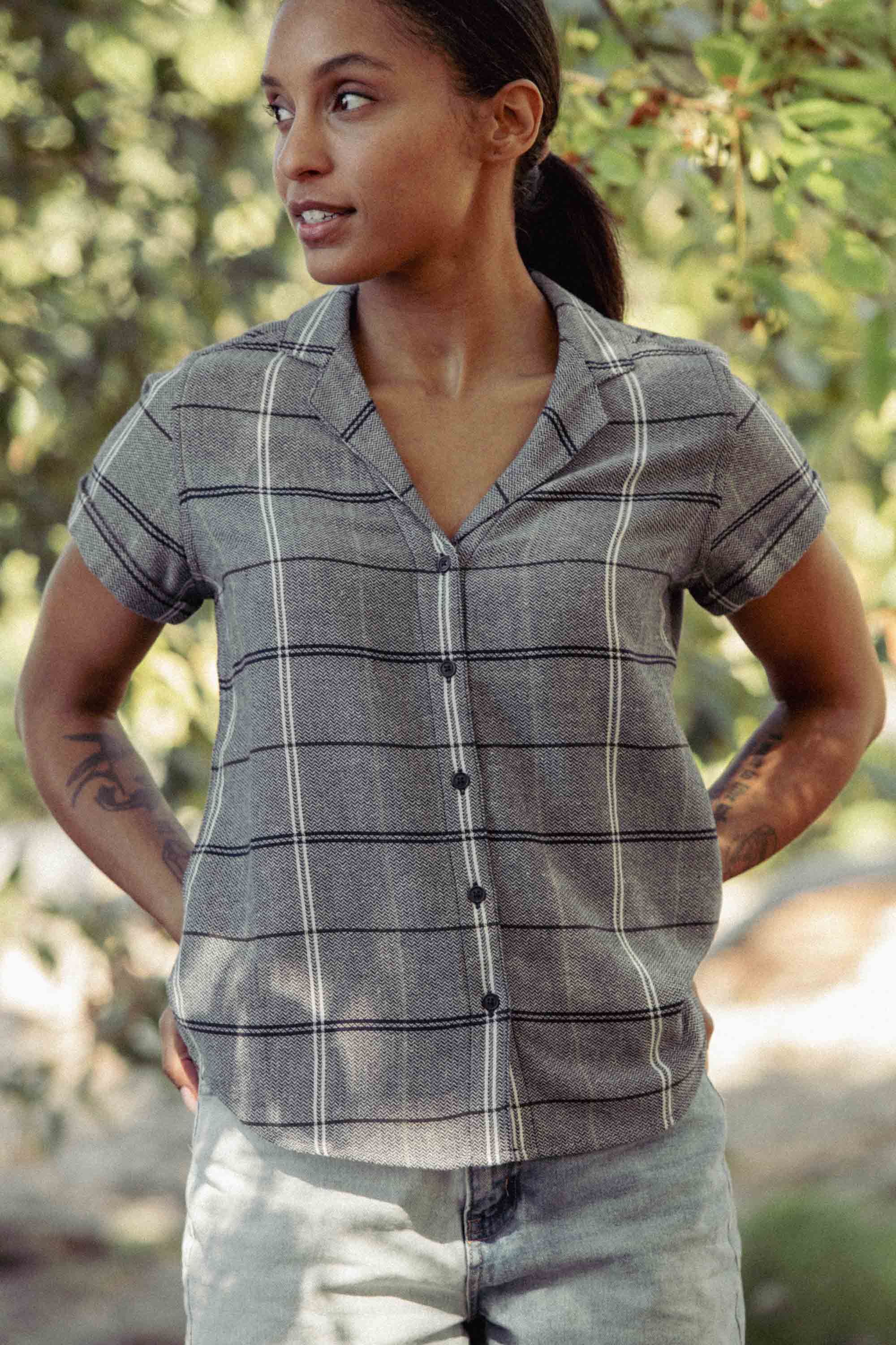 A person with dark hair tied back stands outdoors in the Bridge & Burn Innes Shirt / Overcast Grid and light jeans, looking to the side with hands on hips, as blurred greenery fills the background.