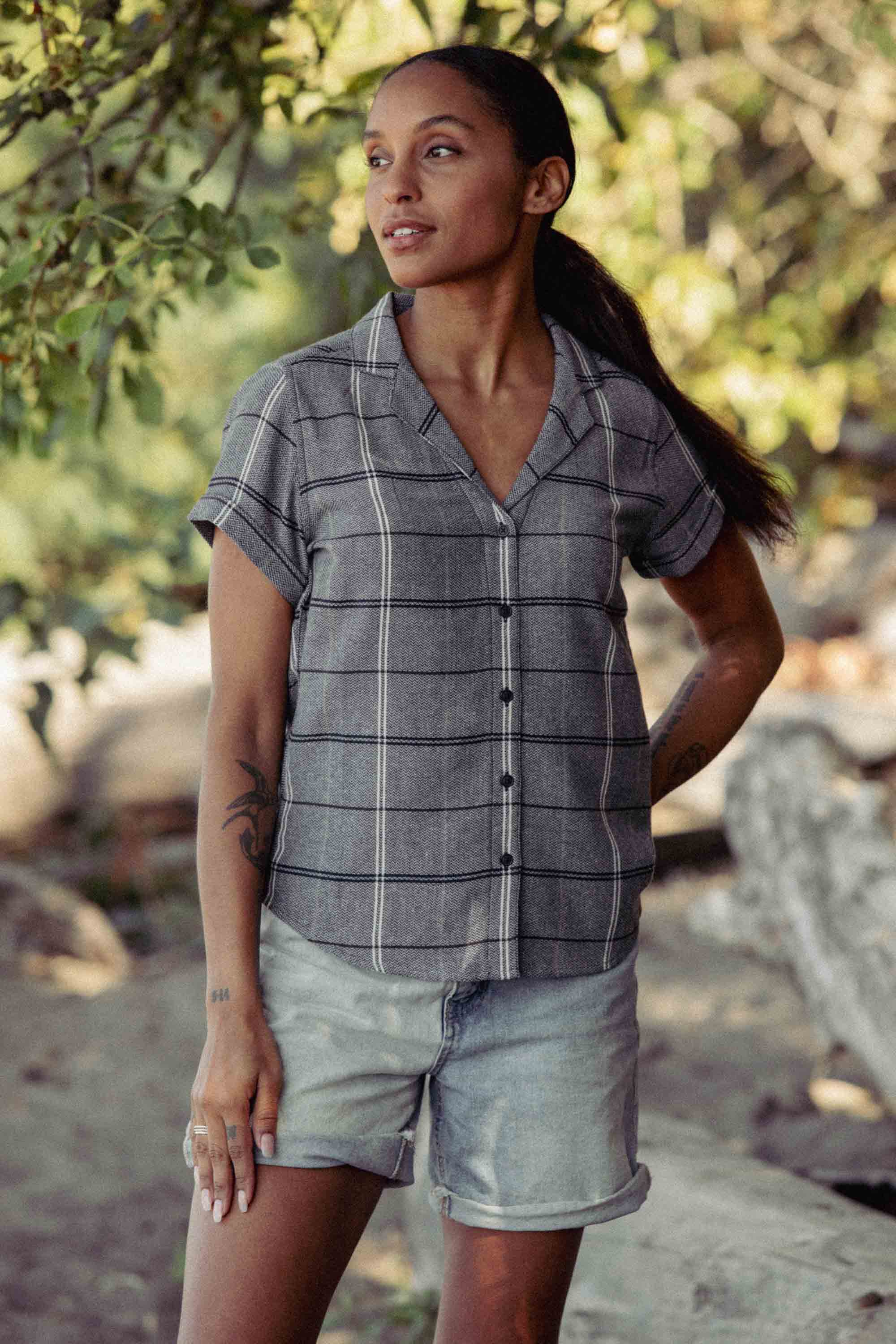 A woman in the Bridge & Burn Innes Shirt in Overcast Grid, a relaxed fit button-up with short sleeves, stands outdoors by trees in sunlight, looking to the side with a calm expression.