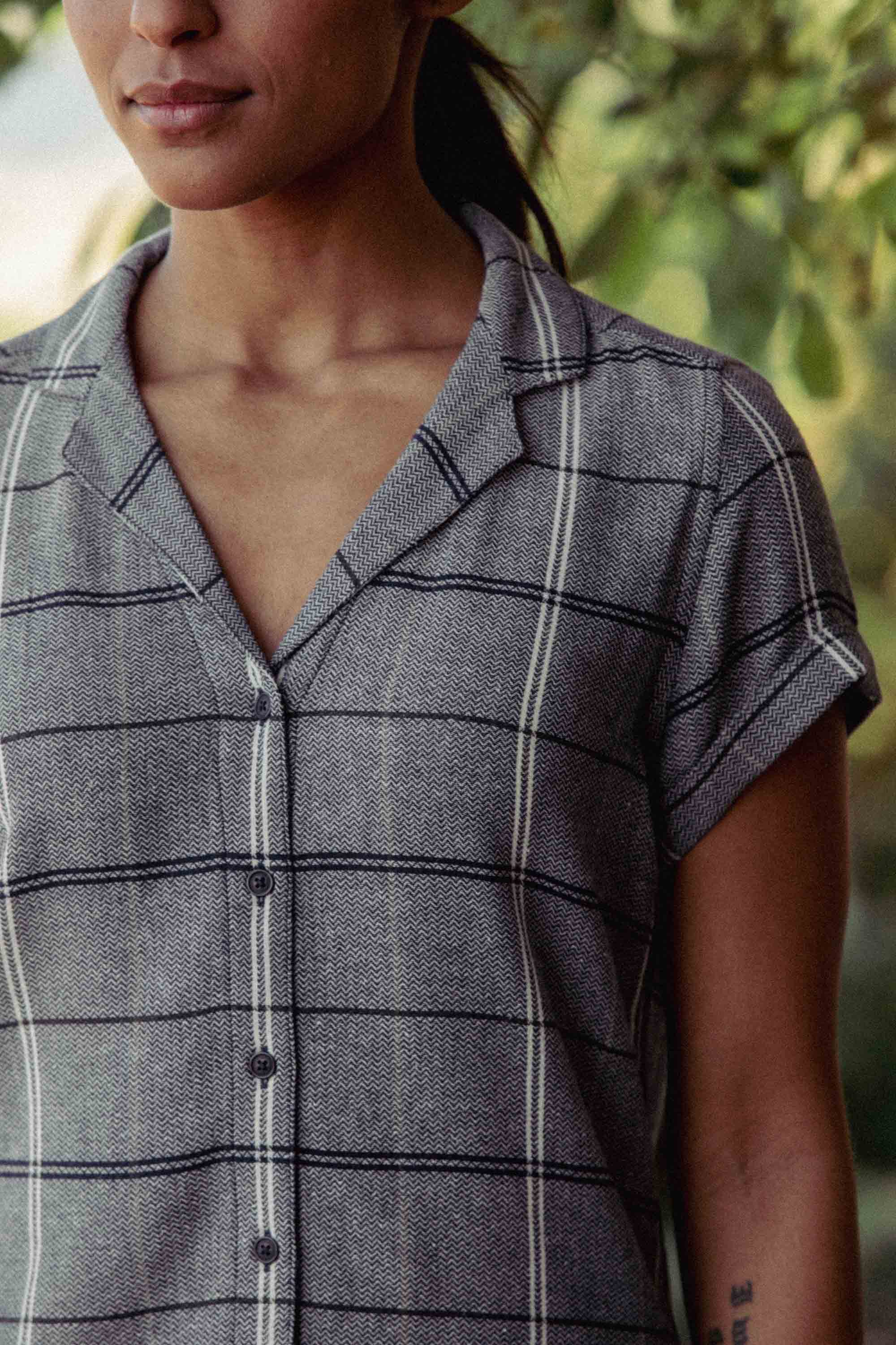 A person stands outdoors near green foliage, wearing the Bridge & Burn Innes Shirt in Overcast Grid—a relaxed fit button-up made from recycled cotton. Their face is partially visible, and their hair is pulled back.