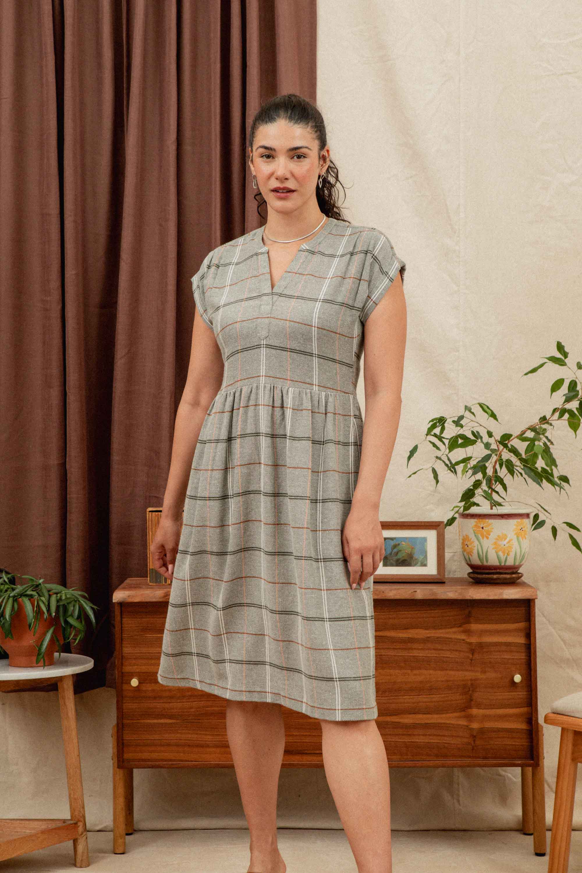 A woman with curly dark hair in a ponytail wears the Bridge & Burn Ira Dress in Olive Ember Grid. She stands indoors near wood furniture, plants, and framed art, with brown curtains and a cream wall in the background.