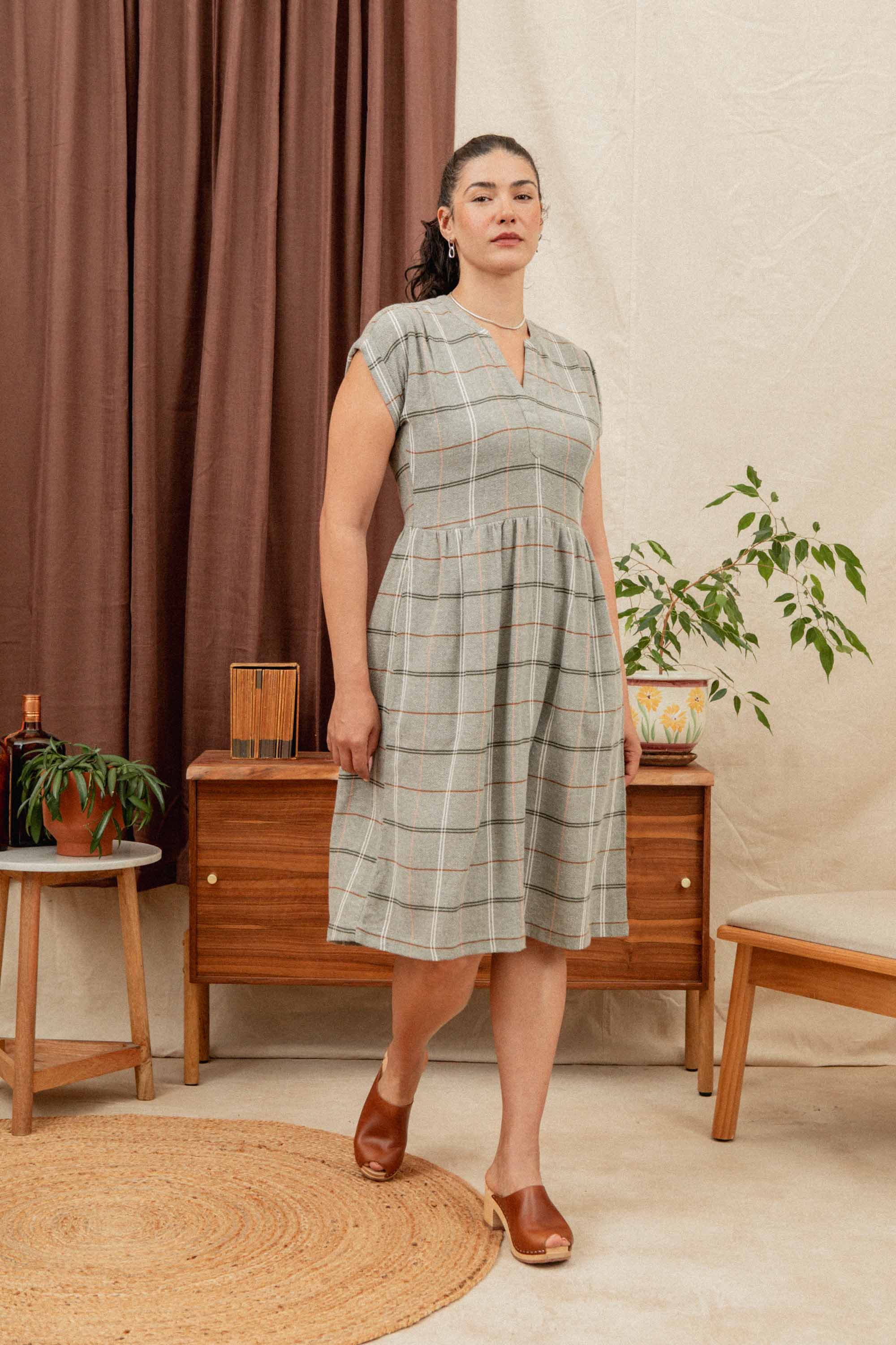 A woman wearing the Bridge & Burn Ira Dress in Olive Ember Grid stands indoors on a circular rug, with wooden furniture, a plant, and brown-and-cream curtains behind her.
