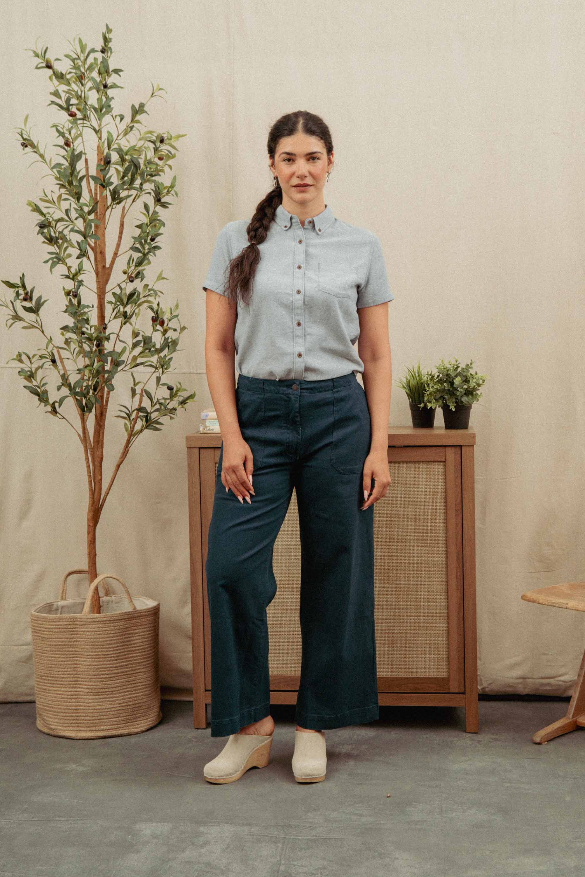 A woman with a braid stands indoors wearing the relaxed fit Bridge & Burn Juniper Shirt in Denim Twill, paired with wide-leg recycled cotton pants. Behind her are a wooden cabinet, small plants, a wicker basket with a tall plant, and a wooden chair.