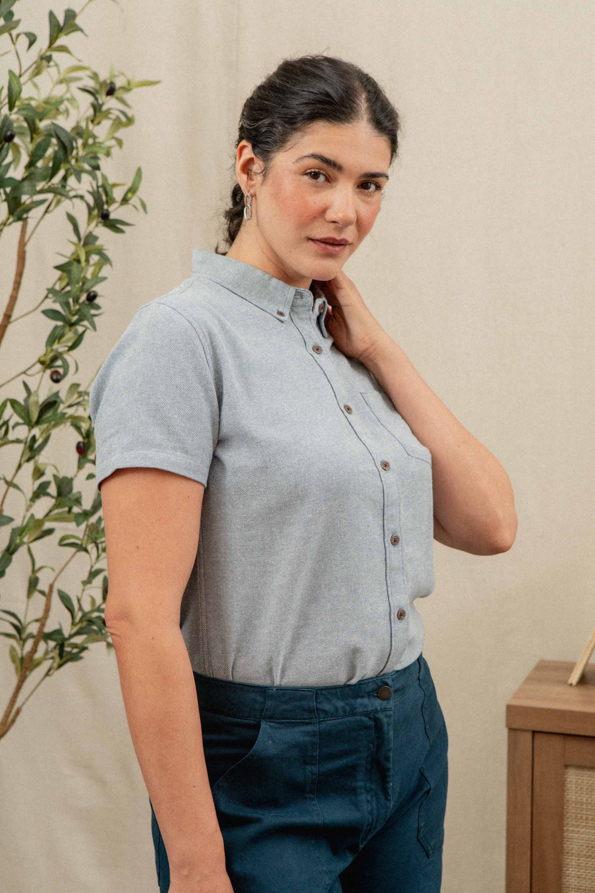 A woman with dark braided hair stands indoors near a plant and wood furniture, wearing the Bridge & Burn Juniper Shirt in Denim Twill and blue pants, looking slightly to the side.