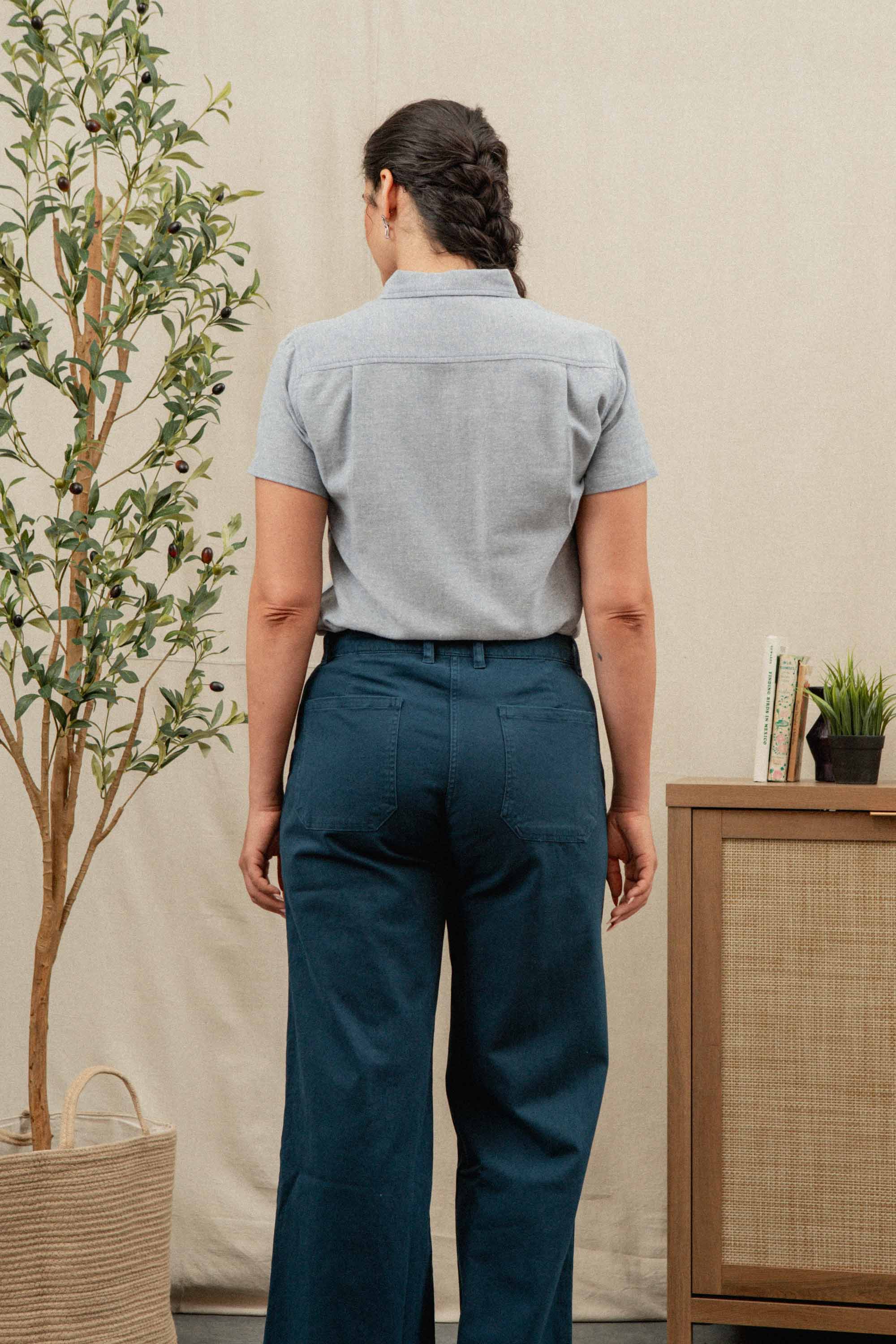 A woman with dark braided hair wears the Bridge & Burn Juniper Shirt in Denim Twill, paired with teal pants. She stands facing away in a room featuring a potted plant and a wooden cabinet adorned with books and decor.
