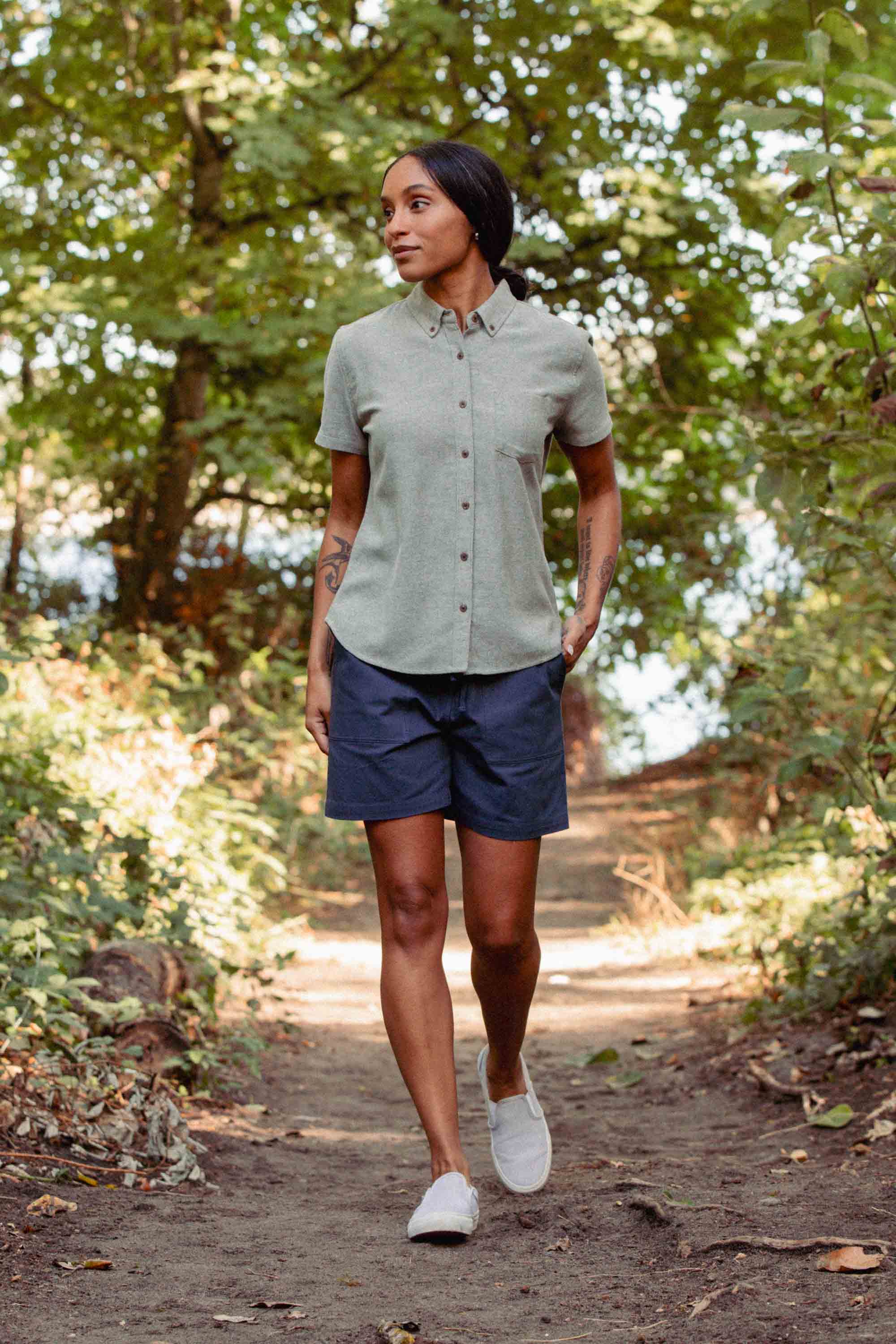 A woman walks on a forest path, wearing the Bridge & Burn Juniper Shirt in Olive Twill with dark shorts. She has tattoos on her arms and hands in her pockets as sunlight filters through the trees.