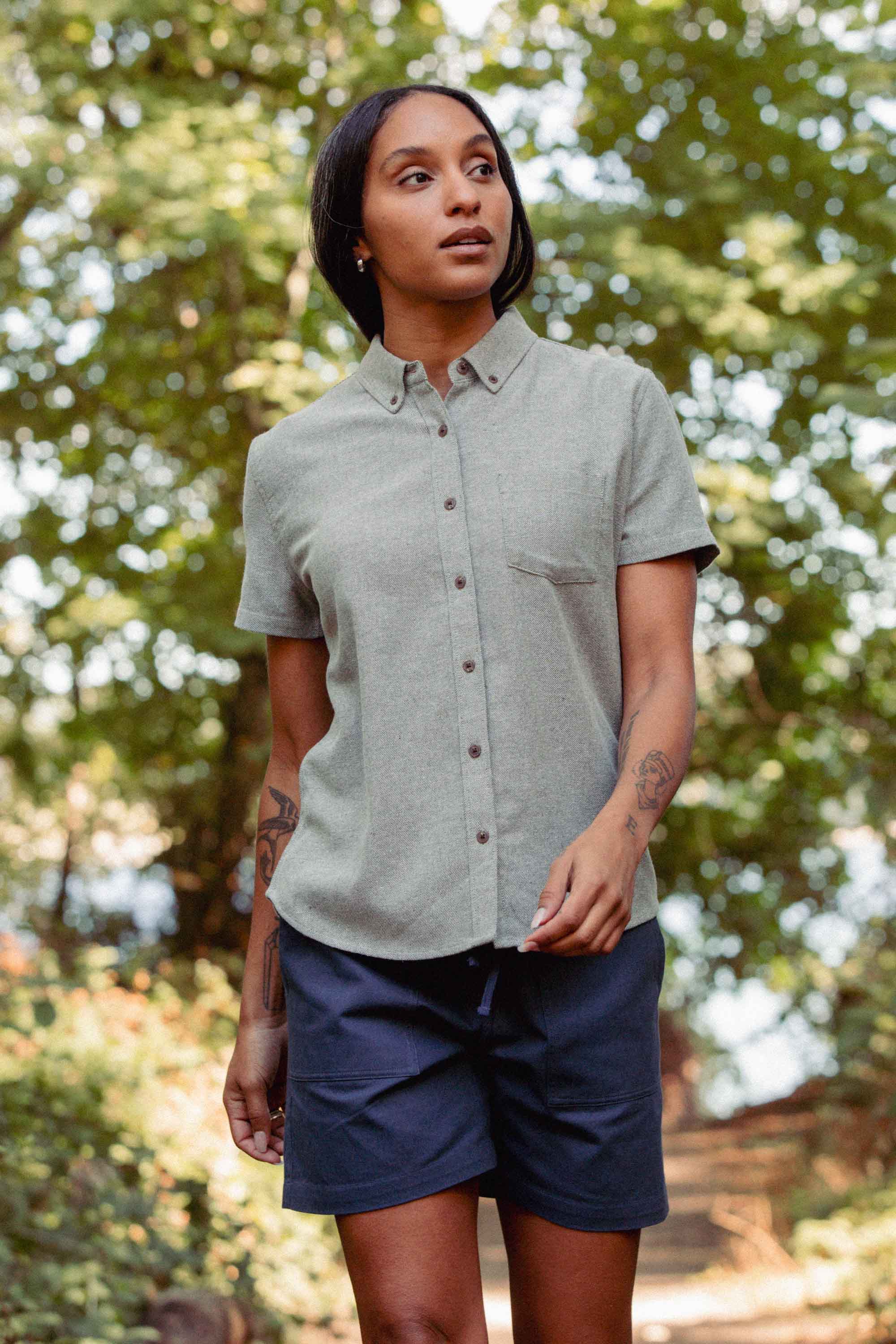 A tattooed individual wearing the Bridge & Burn Juniper Shirt in Olive Twill and navy shorts stands outdoors among green trees, gazing slightly to the side.