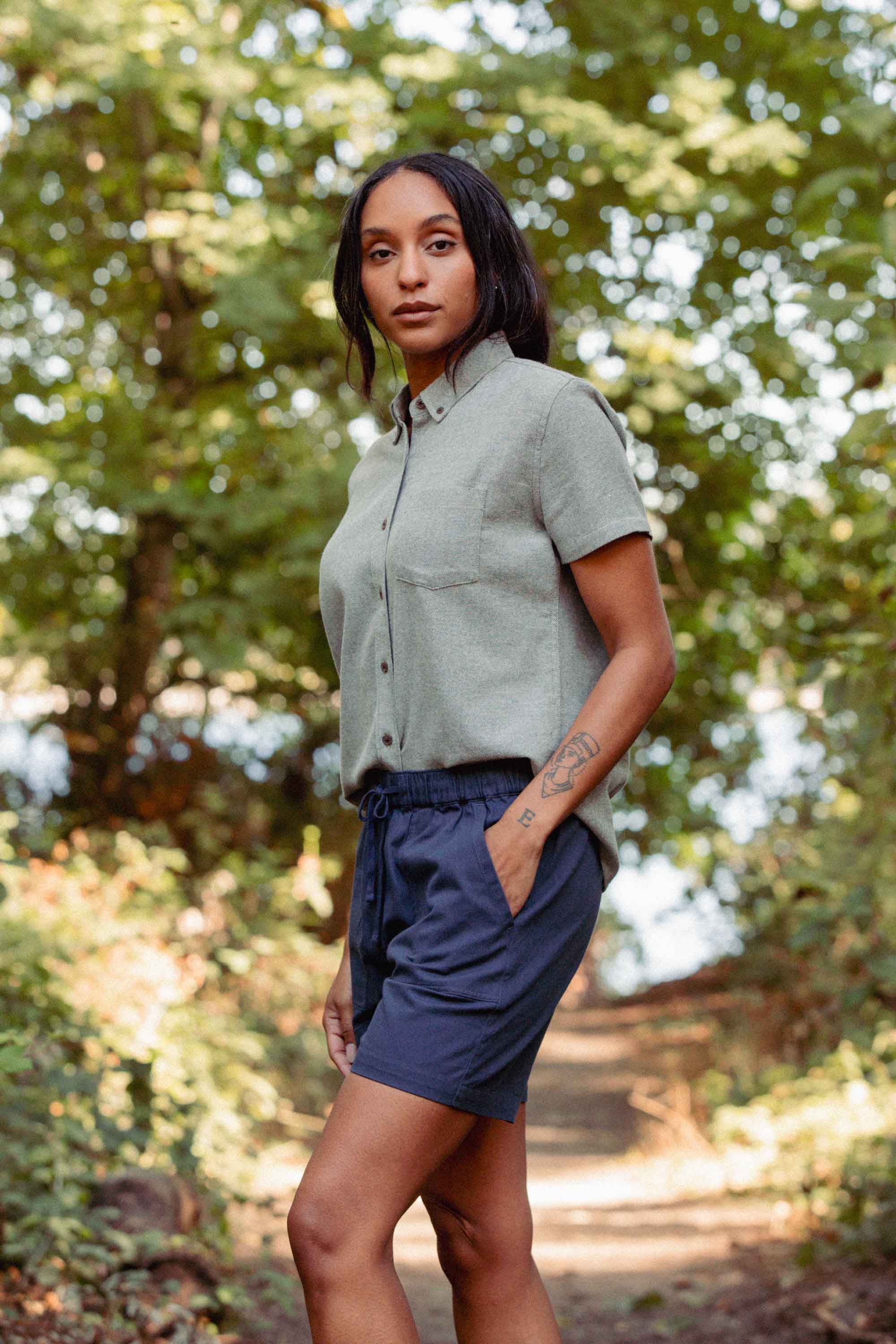 A woman stands on a wooded path wearing the Bridge & Burn Juniper Shirt in Olive Twill and navy shorts, looking calmly at the camera amid lush green trees.