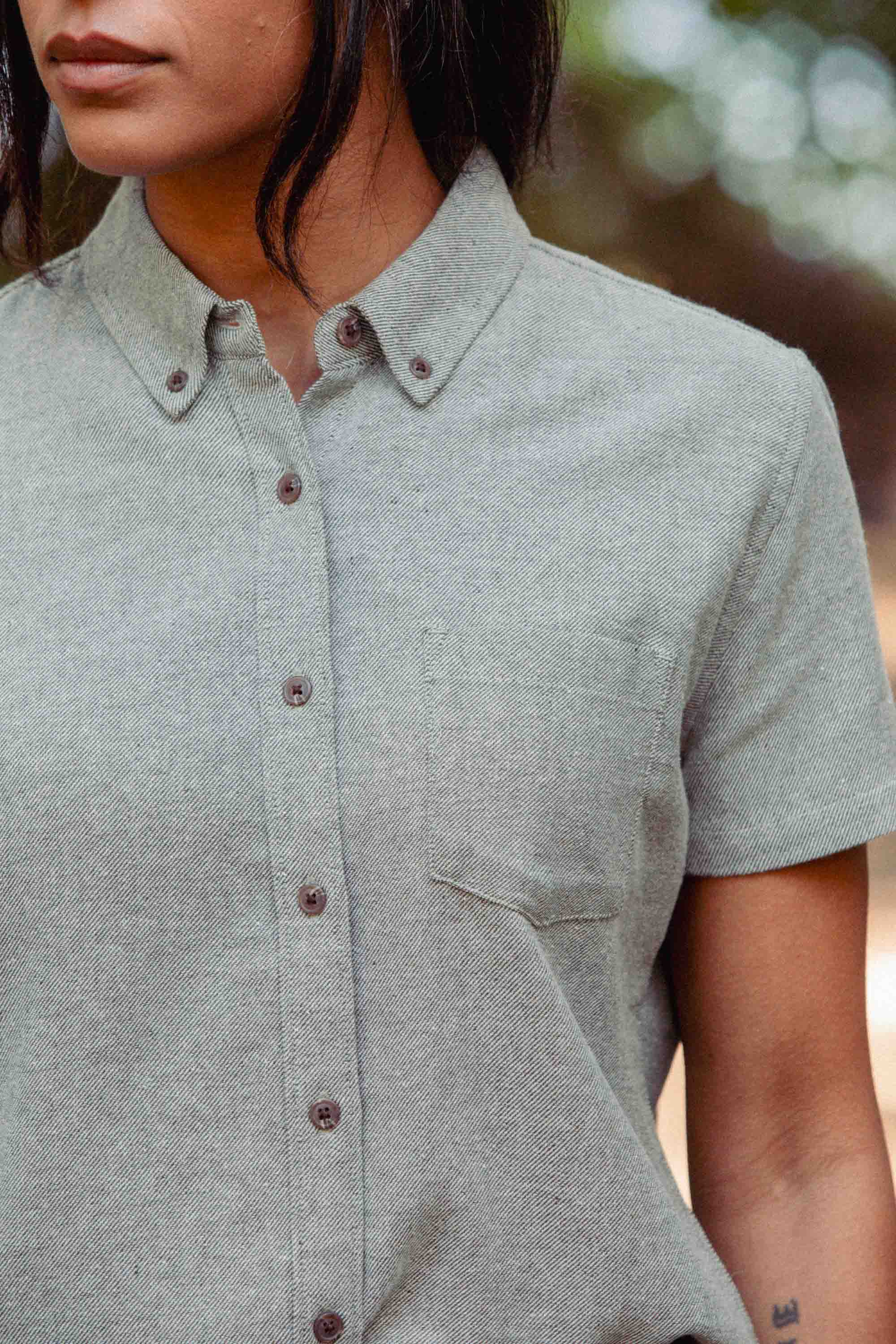 A person in the Bridge & Burn Juniper Shirt in Olive Twill, a relaxed fit short sleeve button-down with a small chest pocket, stands outdoors against blurred green and brown tones. The person's face is partially visible.