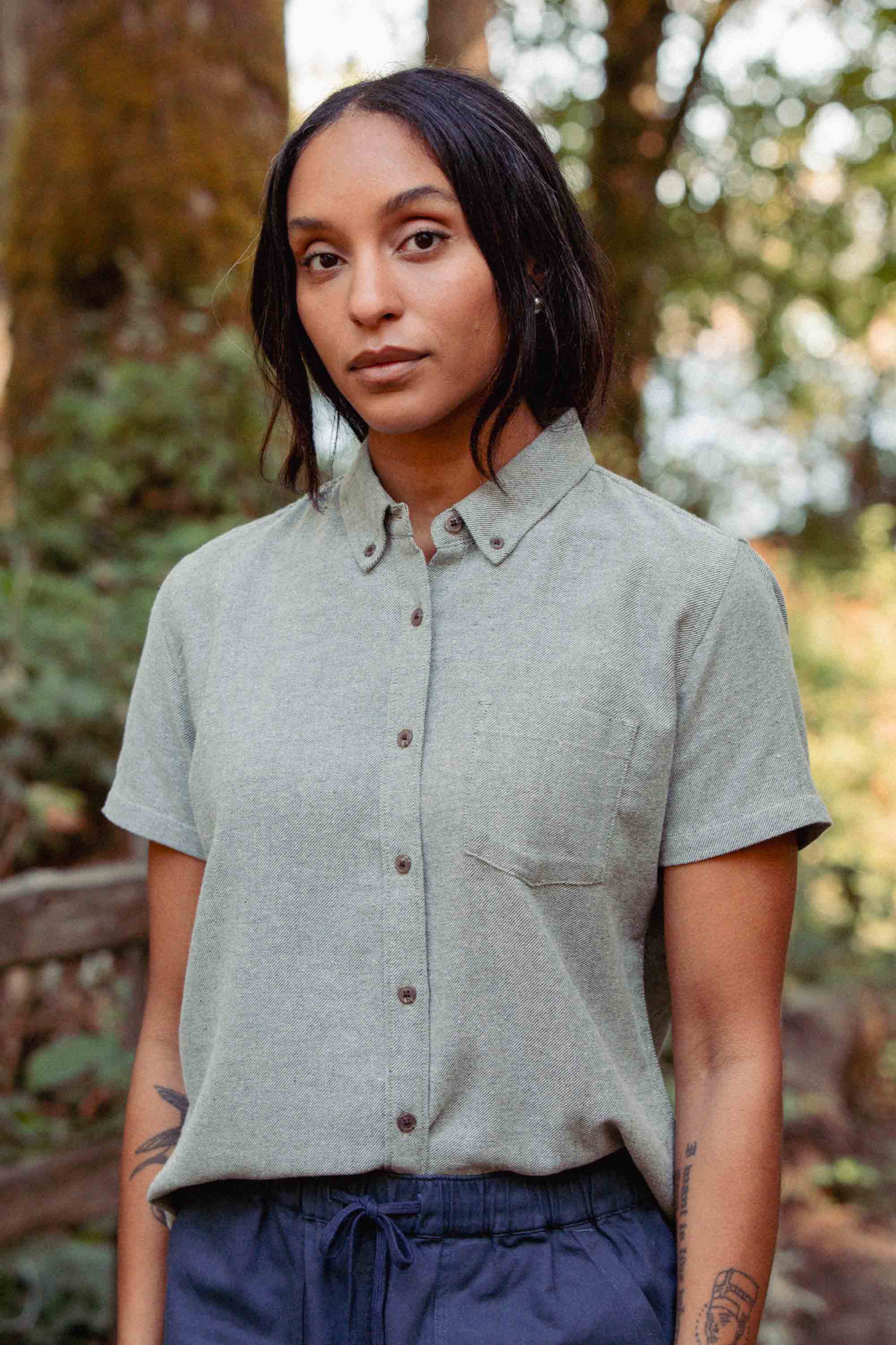 A woman with long dark hair wears the Bridge & Burn Juniper Shirt in Olive Twill and navy pants, standing outdoors in a wooded area and looking at the camera with a neutral expression.