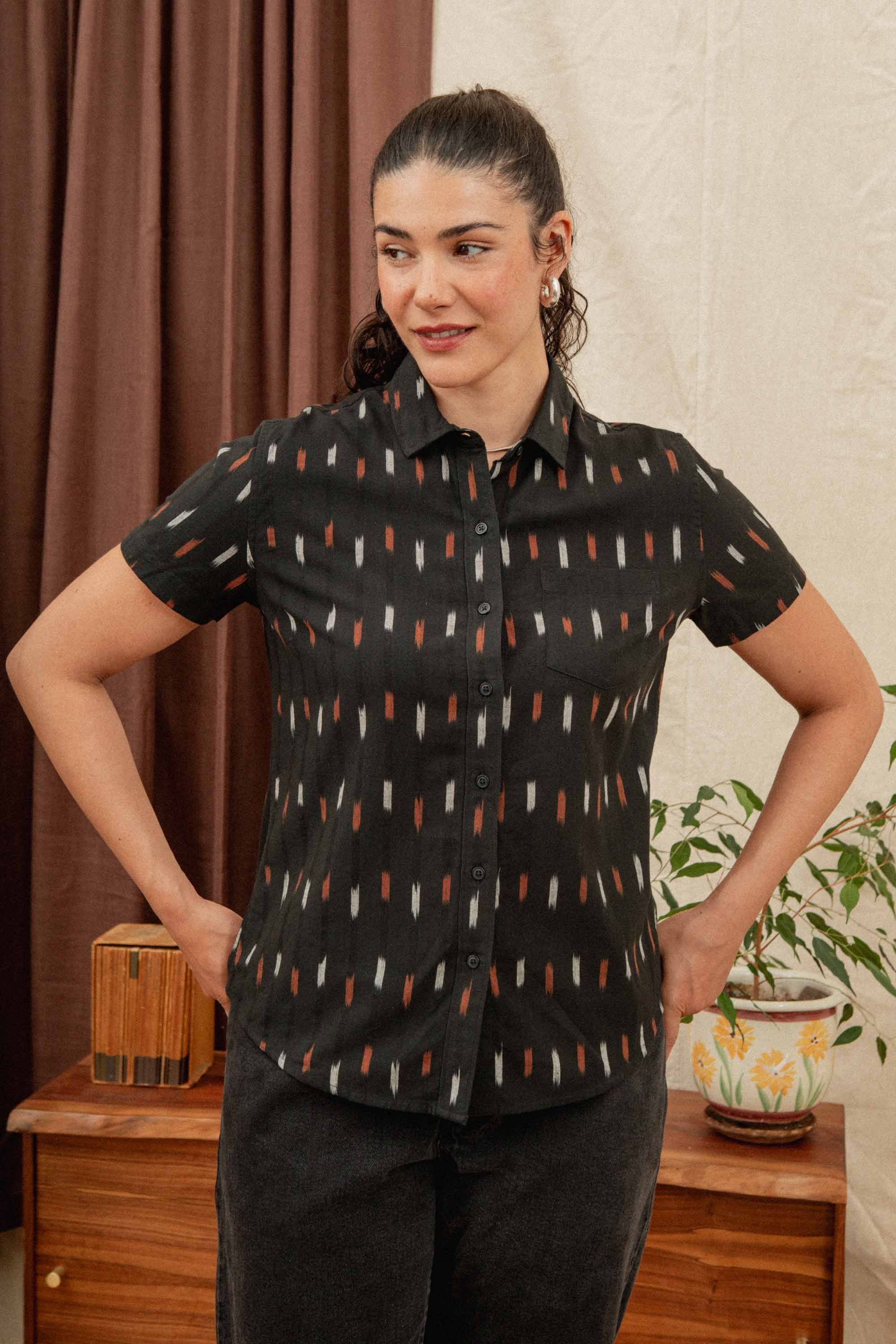 A woman with dark curly hair in a ponytail stands indoors with hands on hips, wearing the Bridge & Burn Lana Shirt in Dotted Horizon—a relaxed fit, 100% cotton black short sleeve button-up with a geometric pattern.