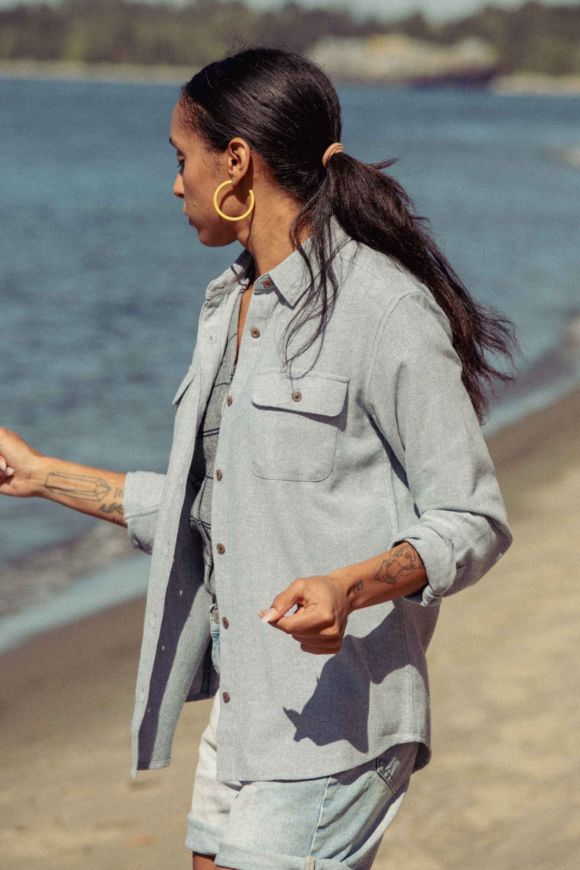 A person with long dark hair in a ponytail wears the Bridge & Burn Lenon Shirt in Denim Twill, standing on a sandy beach near the water and looking to the side with one arm slightly raised.