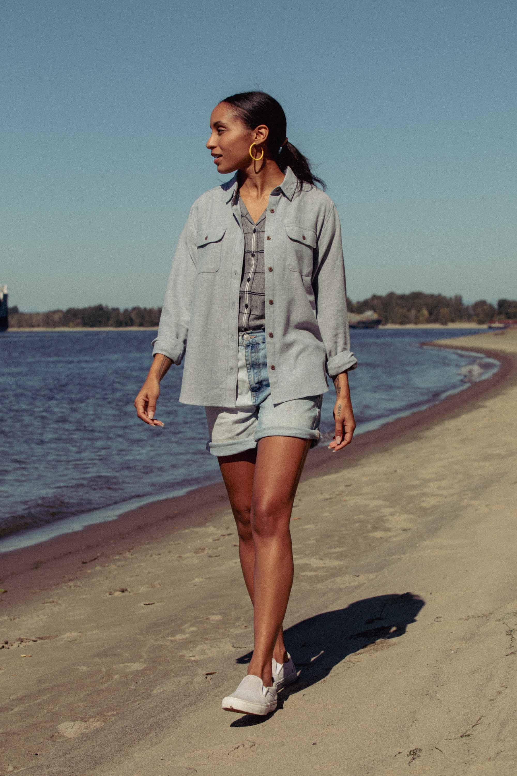 A woman walks along a sunny beach near the water, wearing the Bridge & Burn Lenon Shirt in Denim Twill over denim shorts, with a light denim jacket, white slip-ons, and large hoop earrings. Trees sway gently in the background.
