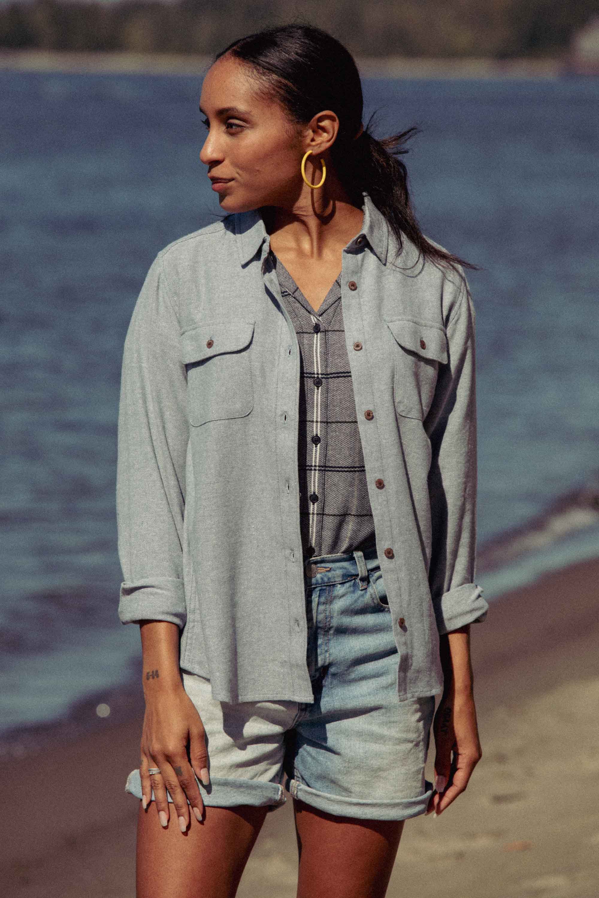 On a sandy beach, a woman wears the Bridge & Burn Lenon Shirt in Denim Twill over a plaid top and denim shorts. Her hair is tied back as she looks to the side, one hand resting by her leg.