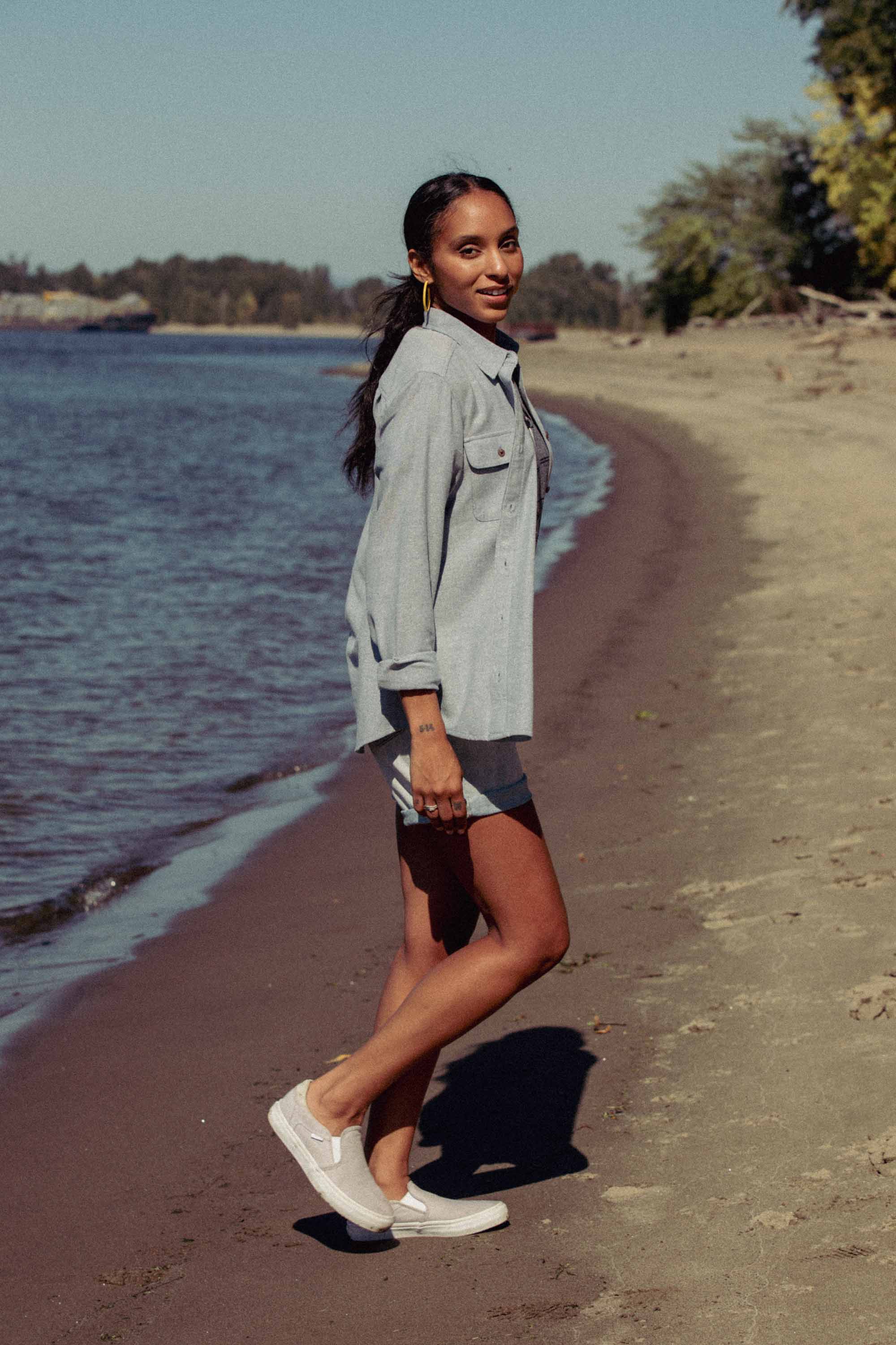 A woman stands on a sandy beach, smiling as she looks back. She wears the Bridge & Burn Lenon Shirt in Denim Twill, matching shorts, and white slip-on shoes. Trees and blue sky complete the background scene.
