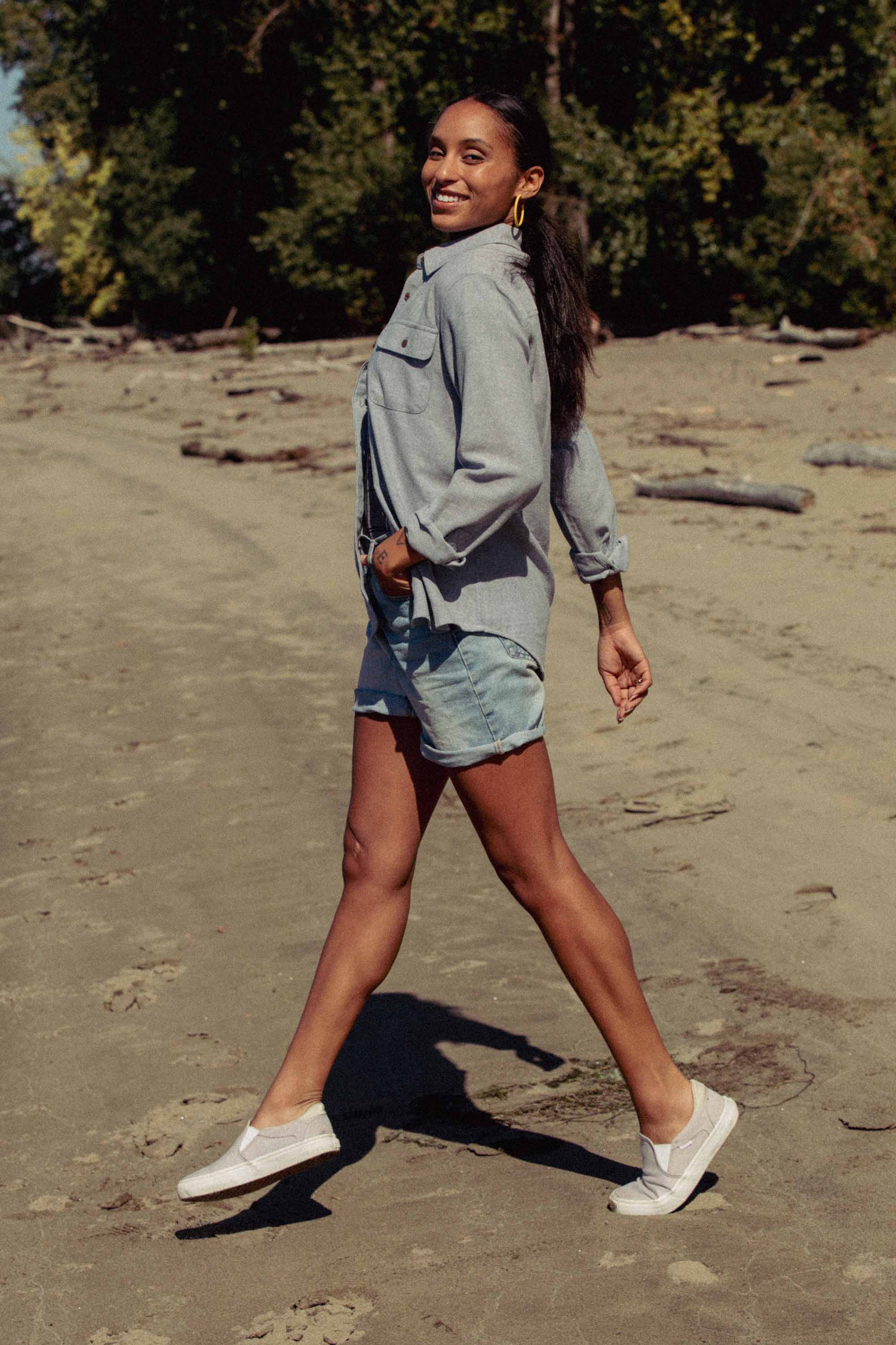 A woman with long hair in a ponytail, wearing the Bridge & Burn Lenon Shirt in Denim Twill, denim shorts, and white sneakers, walks confidently on a sandy beach with trees in the background.