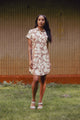 A woman stands on grass in front of a textured wall, wearing the Bridge & Burn Loren Shirt Dress in the Pop Leaf print and light-colored sandals.