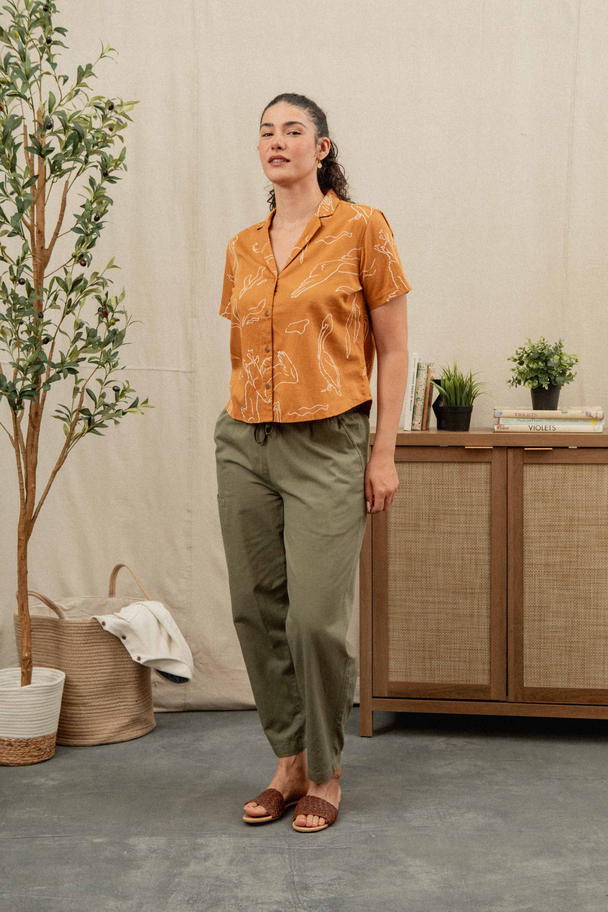 A woman stands indoors beside a plant and sideboard, wearing the Bridge & Burn Mila Cropped Shirt in Shoreflight—an orange organic cotton shirt with a white abstract pattern—paired with olive green pants and brown sandals. Books and plants are in the background.