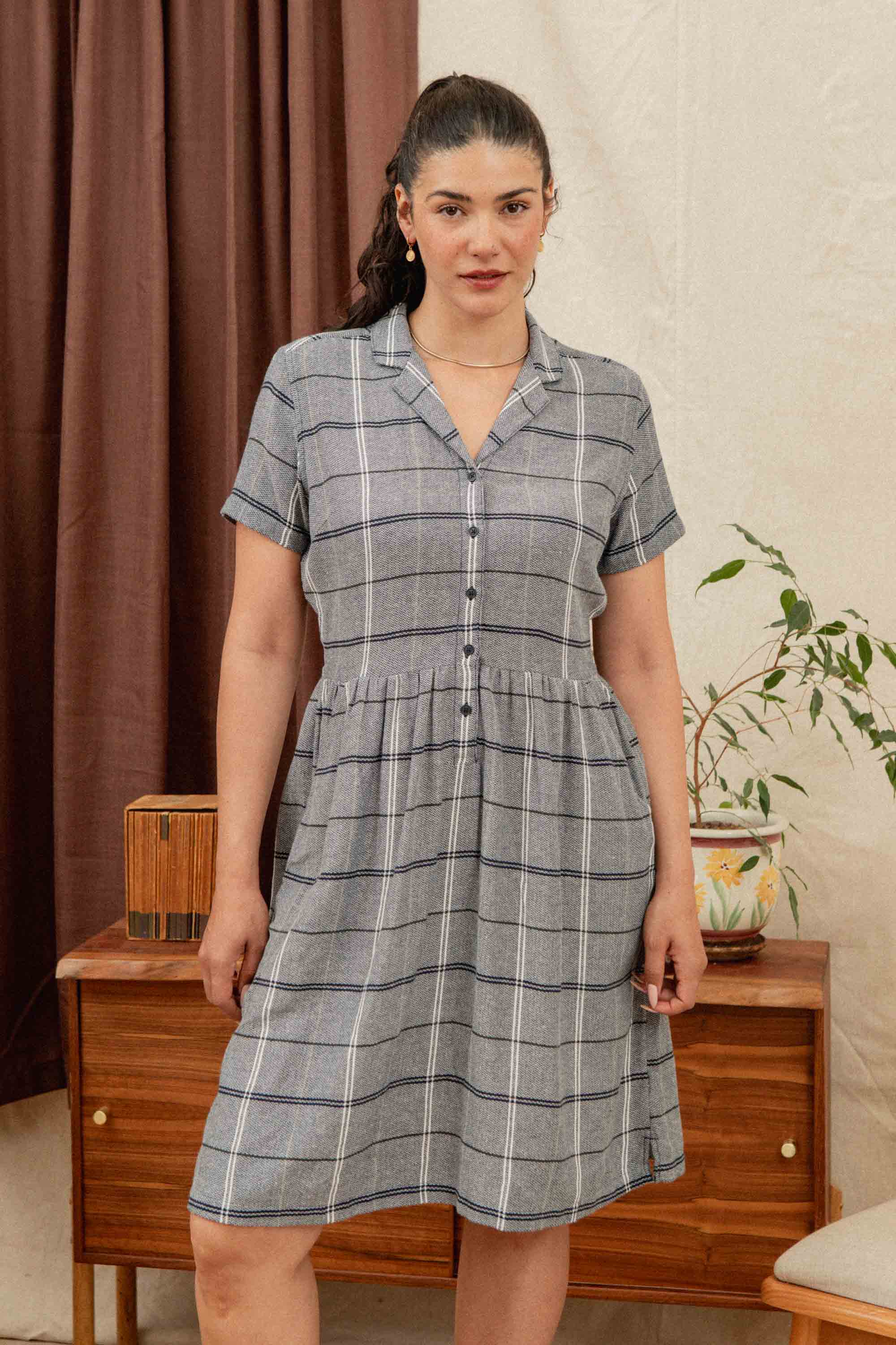 A woman with dark hair in a ponytail wears the Bridge & Burn Ruby Shirt Dress in Overcast Grid. She stands indoors by a wooden dresser with plants, decor, and brown curtains against a cream wall.