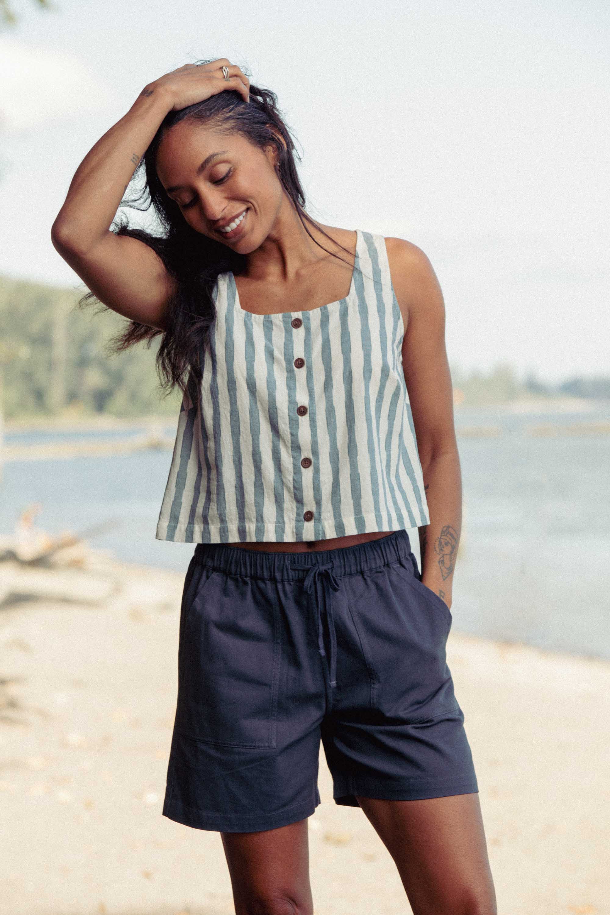 A smiling woman stands outdoors by the water wearing the Bridge & Burn Sadie Tank in Beach Stripe and dark shorts, with one hand in her pocket and the other touching her head. Trees and a blurred shoreline are in the background.