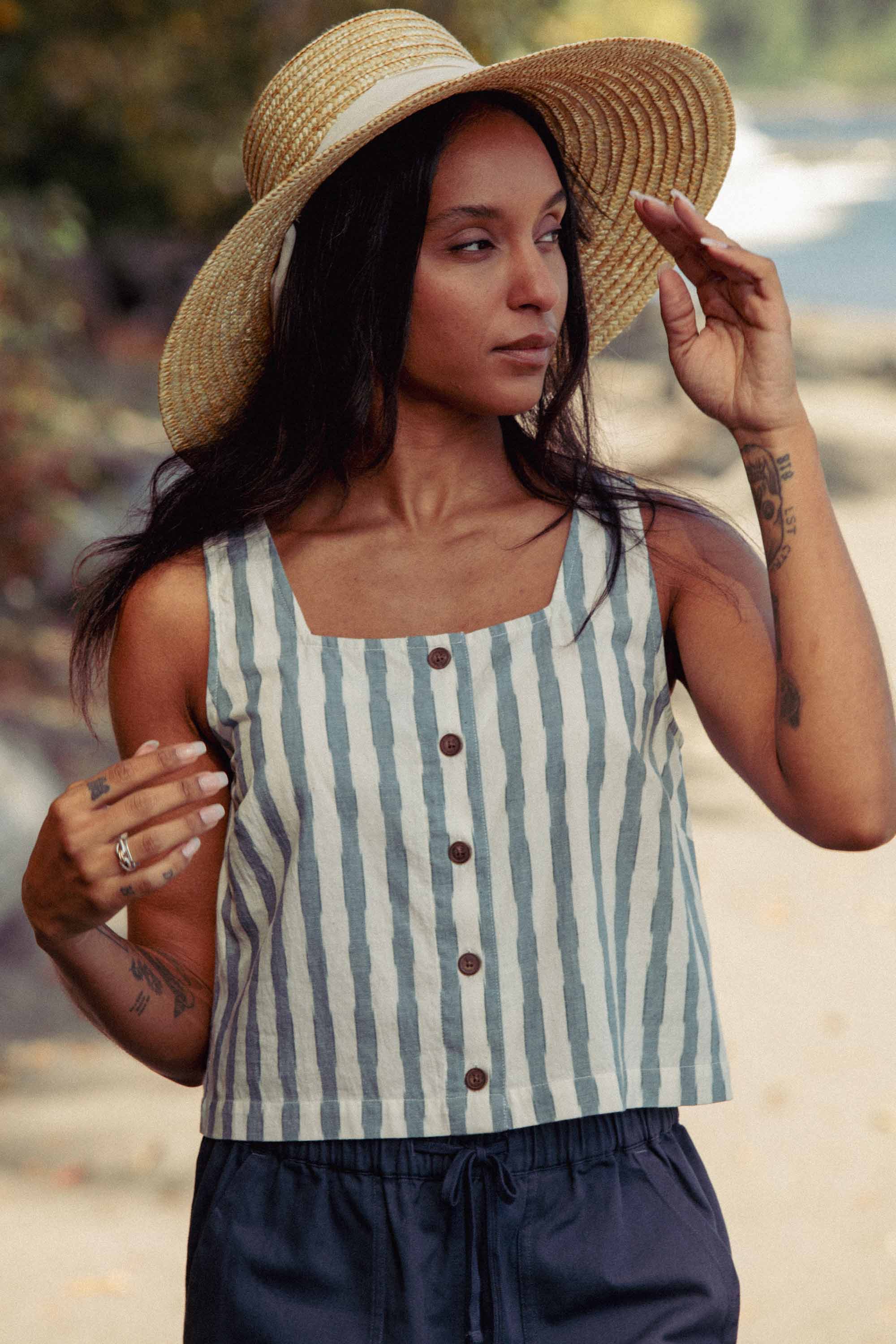 A woman with long dark hair and arm tattoos stands outdoors by trees and water, touching her hat. She wears the Bridge & Burn Sadie Tank in Beach Stripe with dark pants and a wide-brimmed straw hat.